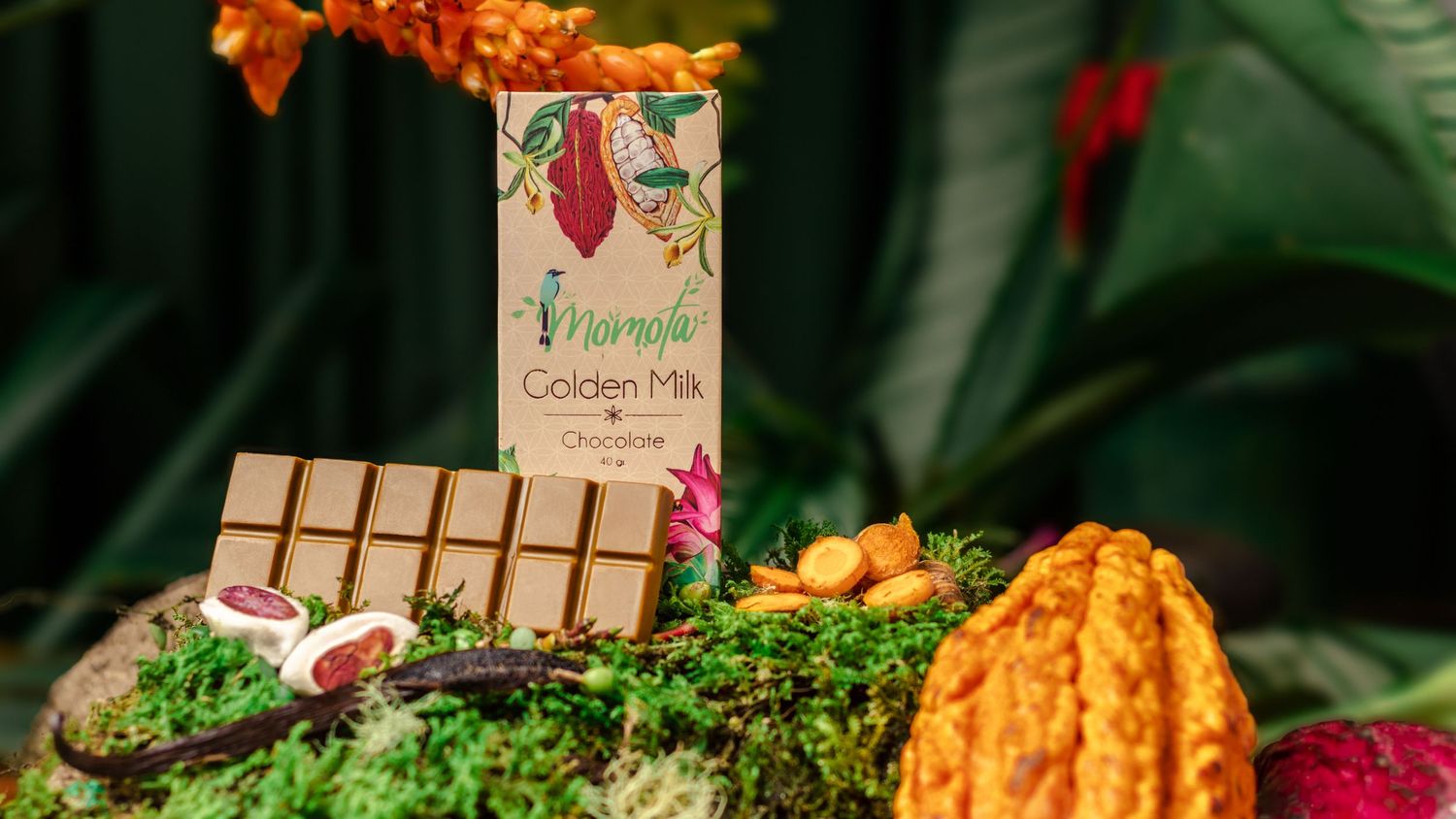 Chocolate bar packaging and cacao pod arranged on moss with tropical flowers.