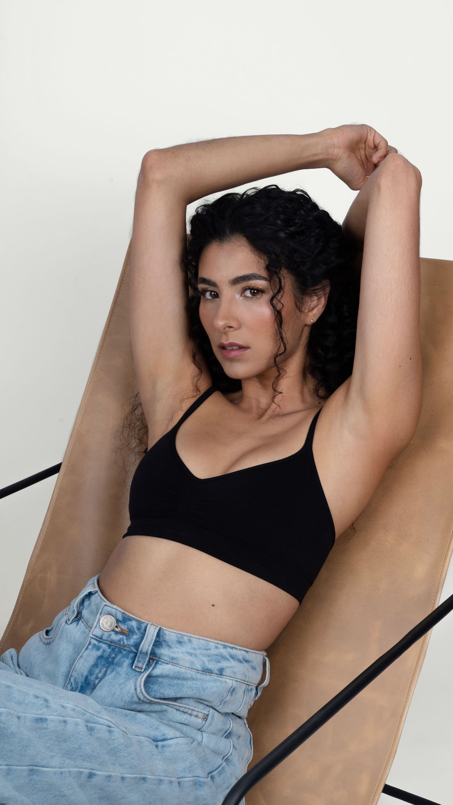 Casual fashion pose in black crop top and light denim against tan chair.