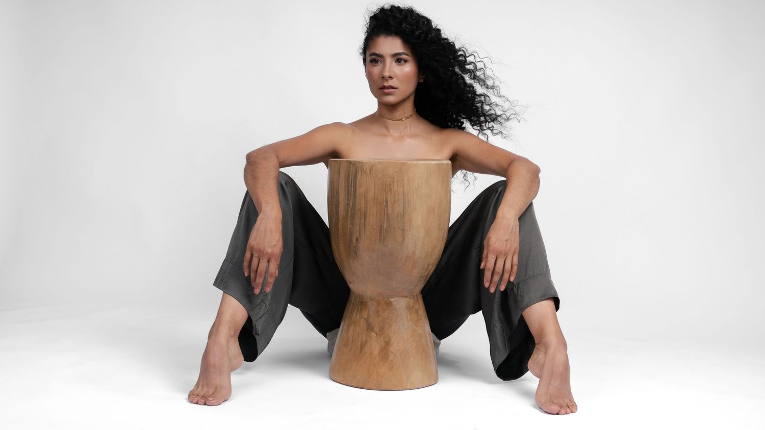 Artistic portrait with model seated on wooden drum stool against white background.