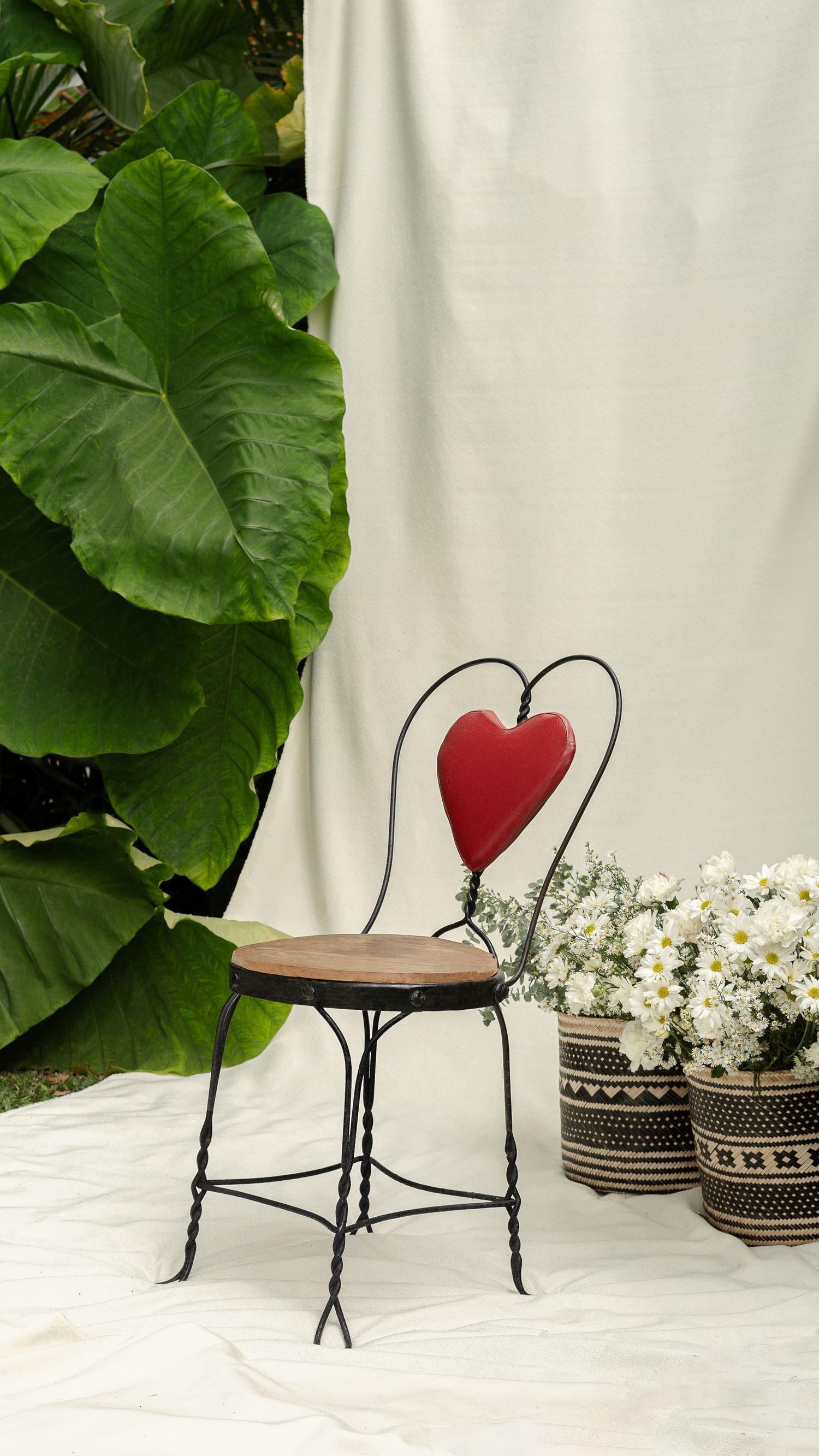 Decorative metal chair with red heart backrest against white backdrop with tropical plants and woven baskets.