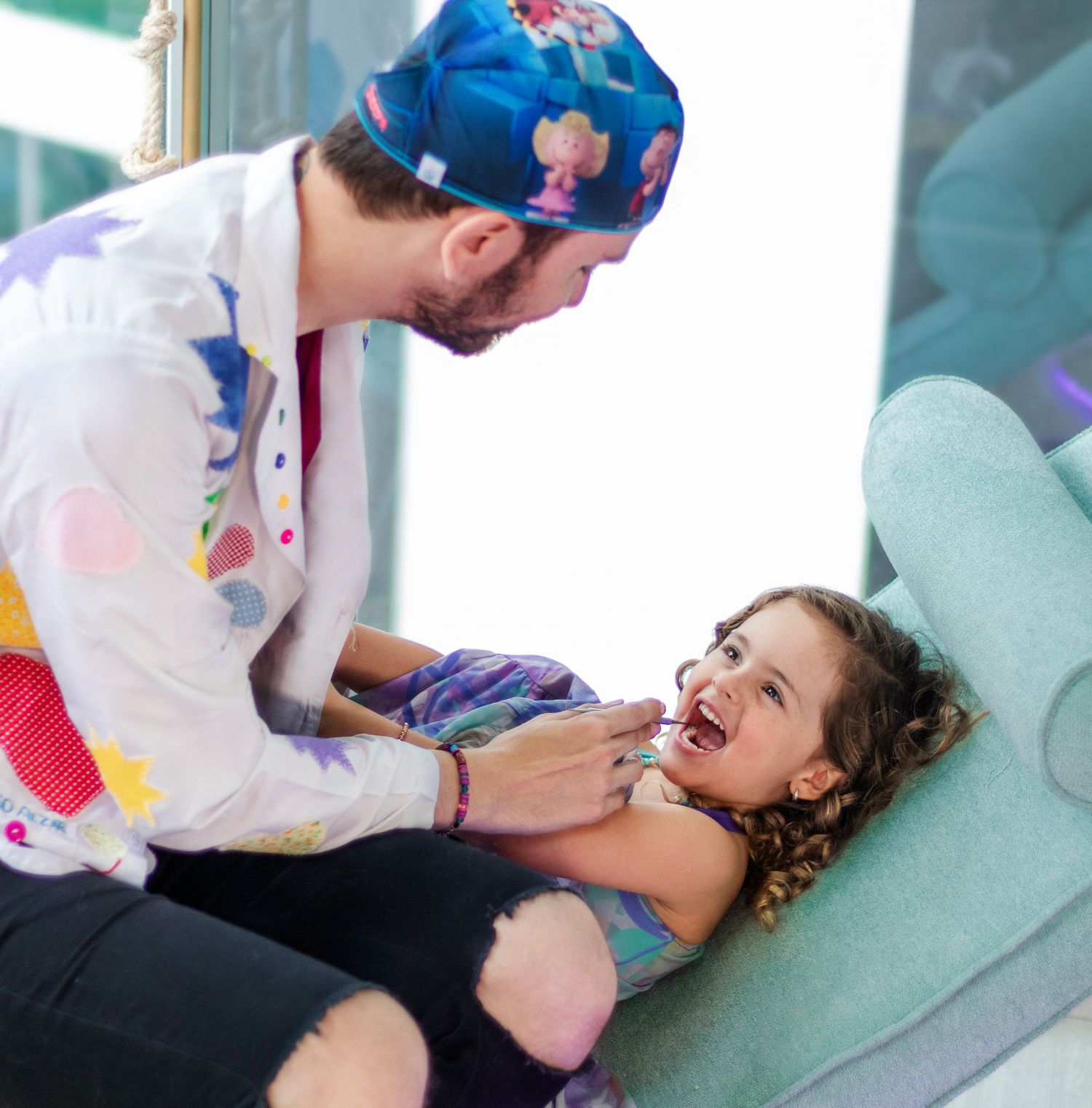 Pediatric dentist wearing colorful scrub cap examines young patient in dental chair.