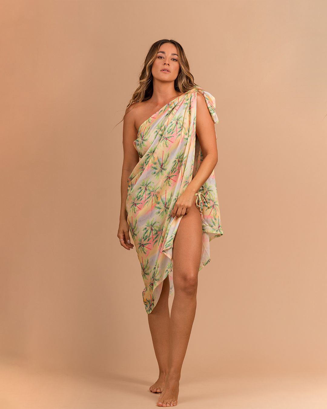 Model posing in a tropical print one-shoulder sarong dress against a beige backdrop.