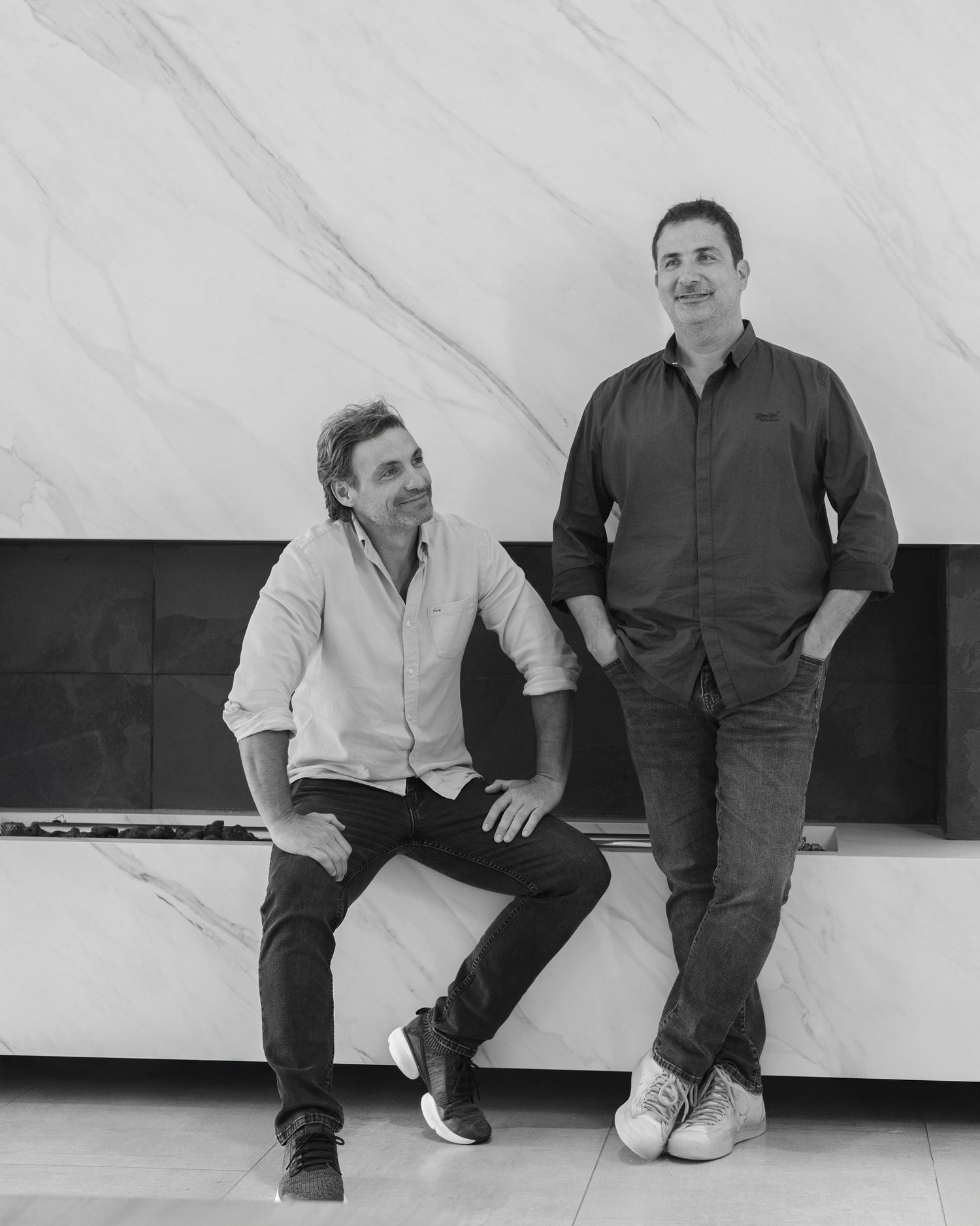 Two men smiling in a modern marble interior, one seated and one standing, in a stylish black and white portrait.