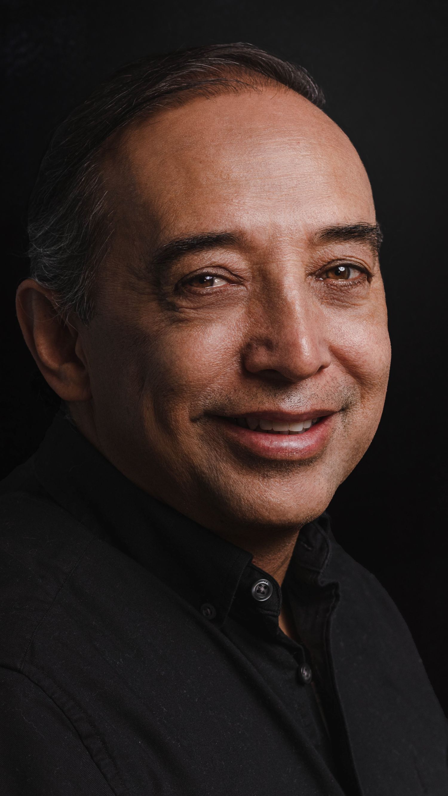 Professional portrait of a smiling man in black attire against dark background.