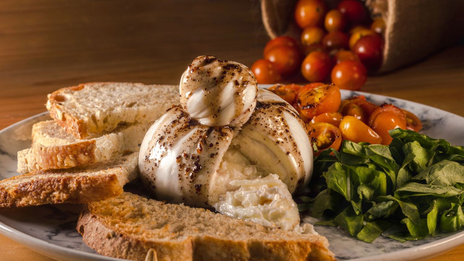Plated burrata cheese with balsamic reduction served alongside bread, tomatoes and fresh greens.