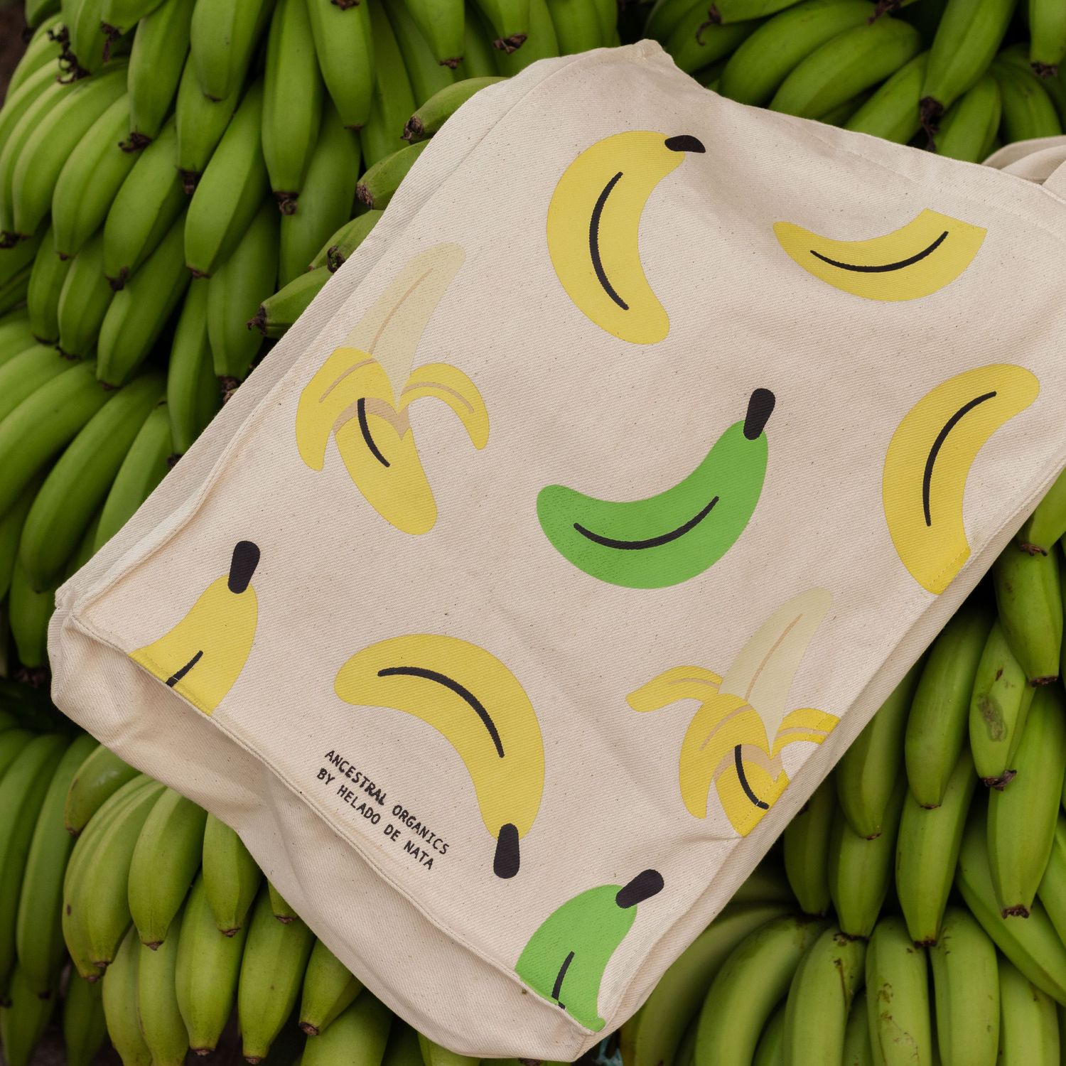 White fabric bag with banana pattern design displayed on fresh green bananas.