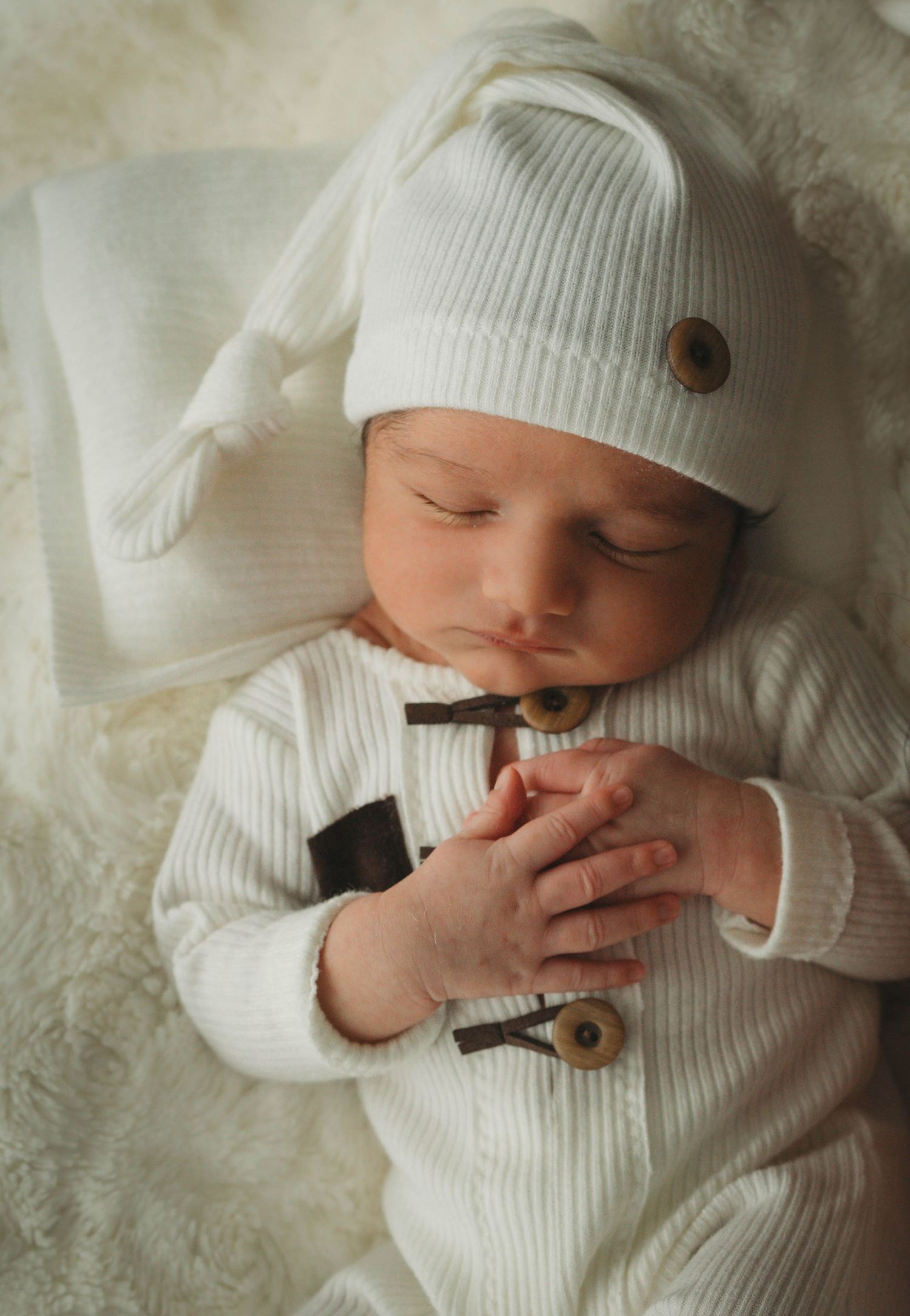 A sleeping baby in white knit clothing and beanie rests on plush white fabric.
