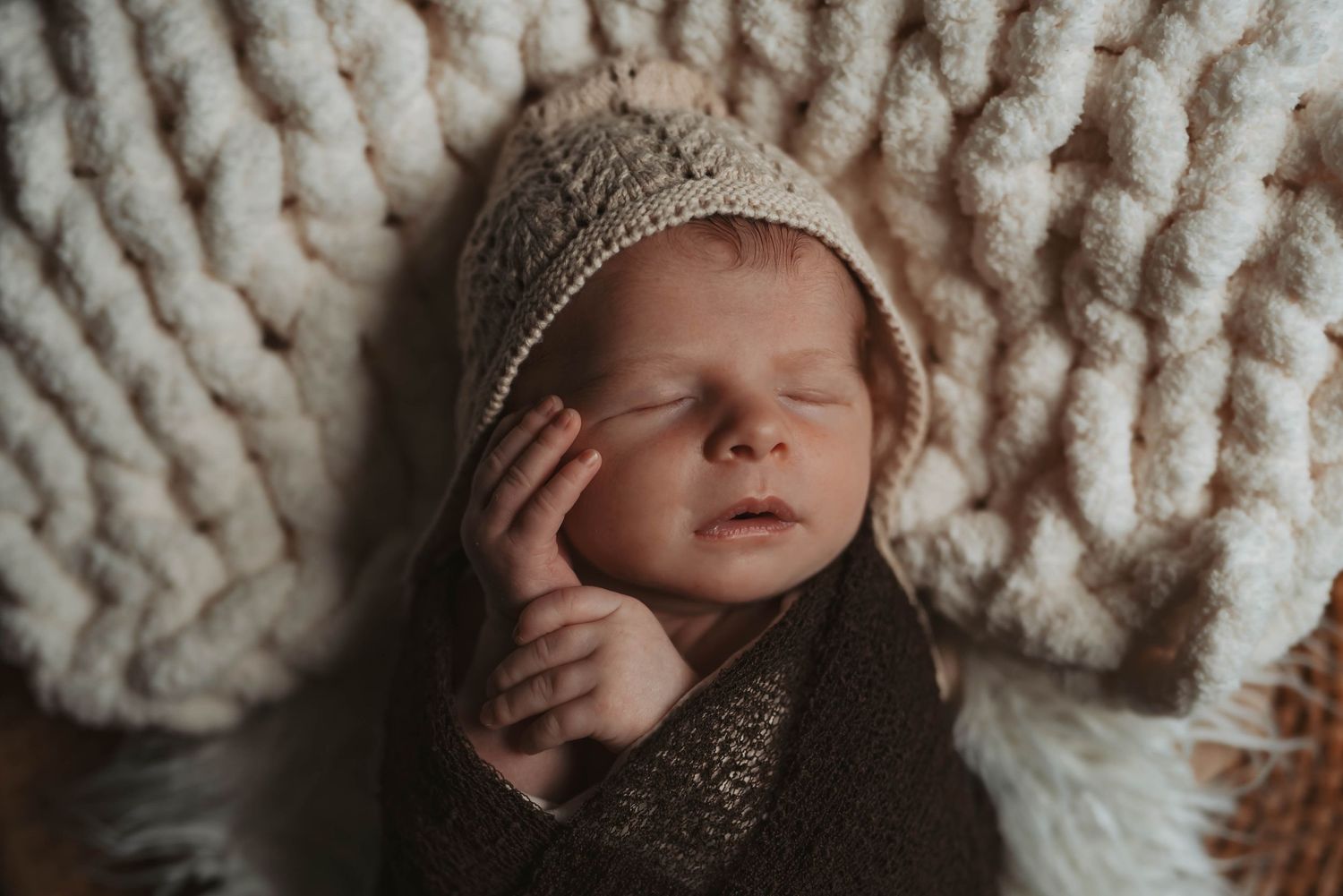Sleeping newborn wrapped in knit blanket on textured white background.
