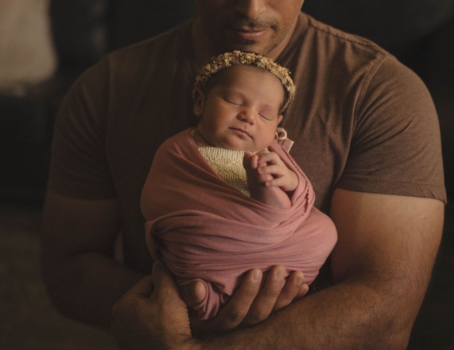 Sleeping newborn in pink wrap and floral crown cradled in strong arms.