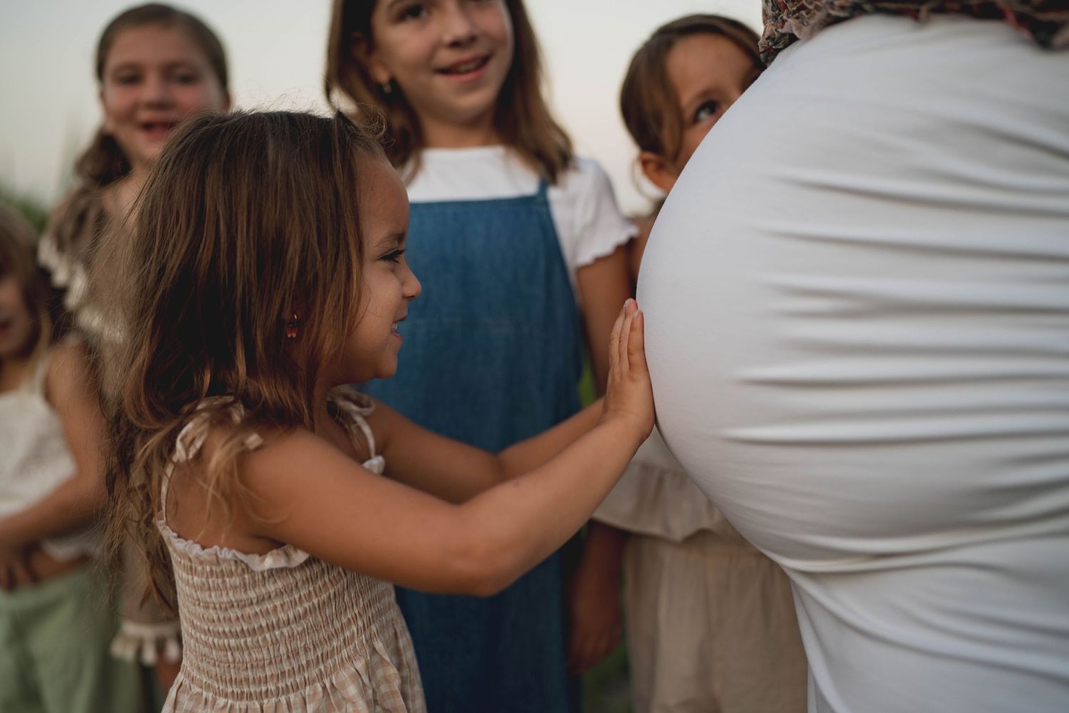 Children gather around to feel a pregnant belly during an intimate outdoor moment.