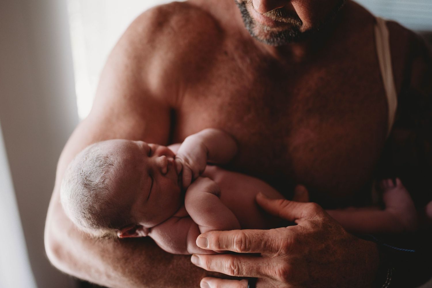 Newborn infant cradled in arms against bare chest in gentle window light.