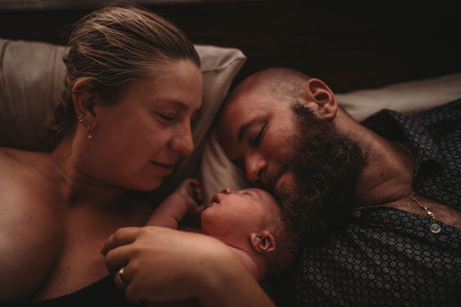 Intimate family moment with newborn baby nestled between parents on bed.