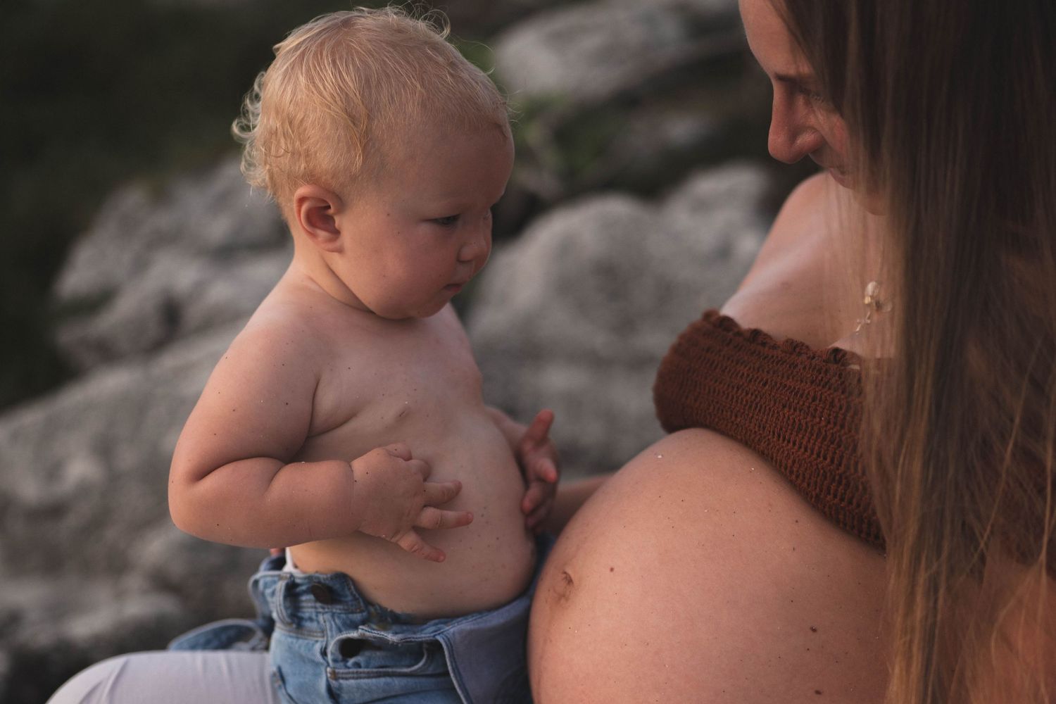 A toddler in denim touches a pregnant belly while sitting outdoors on rocky terrain.