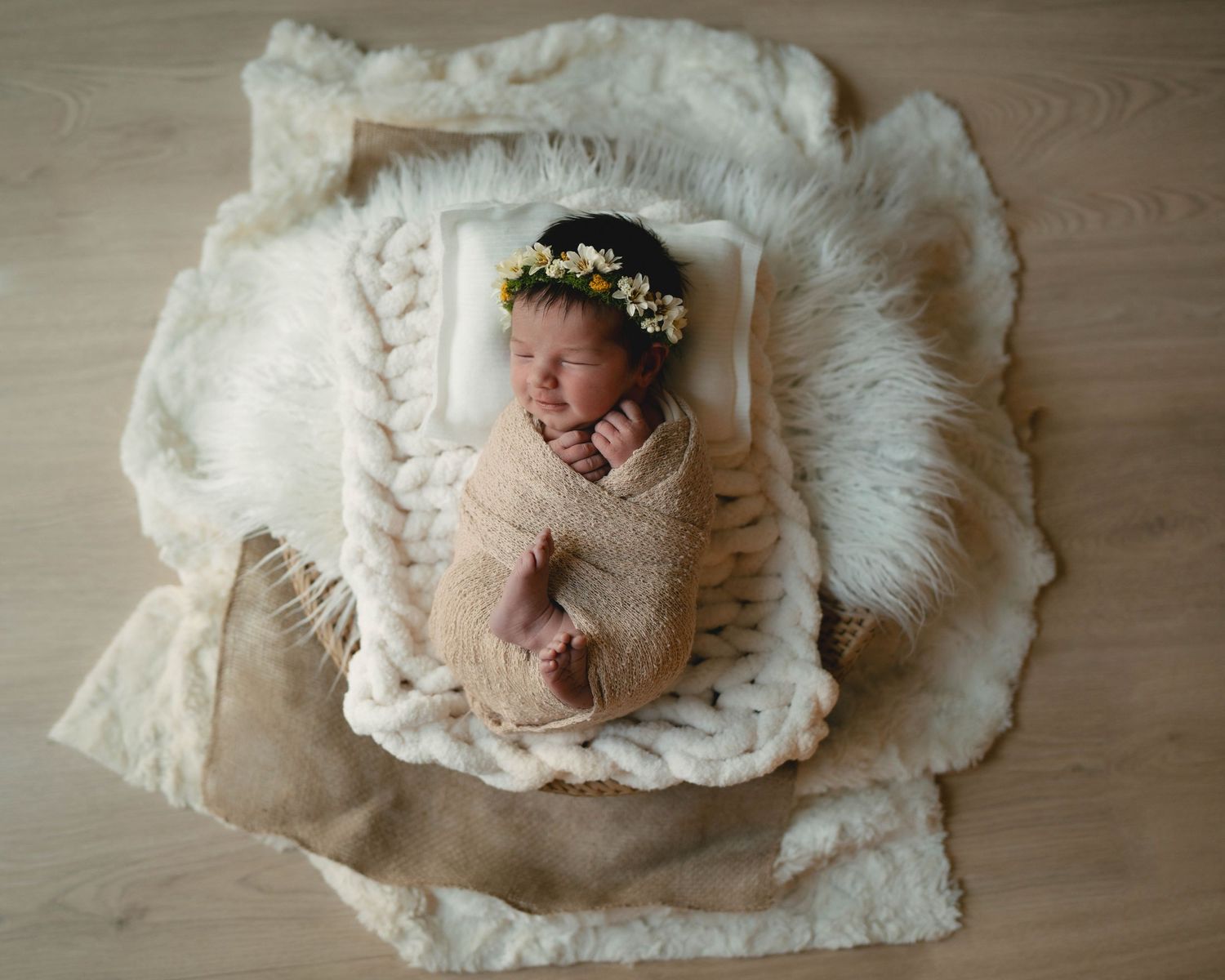 Newborn wearing floral crown sleeps in white basket on textured blankets.