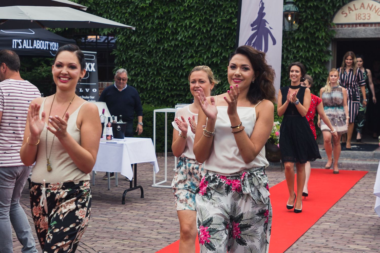 People clapping and celebrating at outdoor event with red carpet and ivy wall.