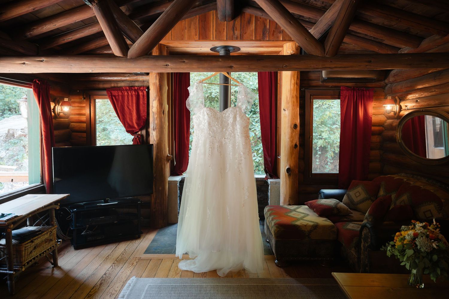 Rustic log cabin interior with exposed wooden beams, red curtains, and large windows overlooking a natural landscape.
