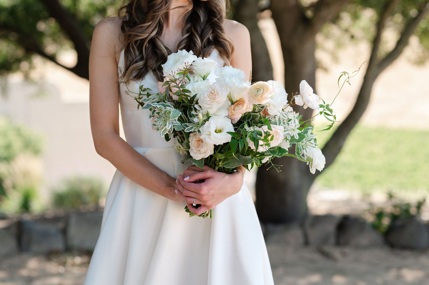 A bride holds a beautiful white and green floral bouquet against her wedding dress in a garden setting.