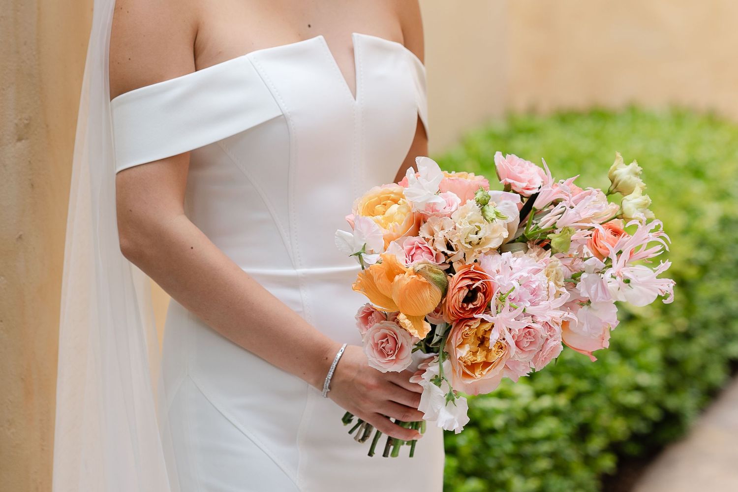 A bride in a white wedding dress holds a peach and pink floral bouquet against a green garden backdrop.