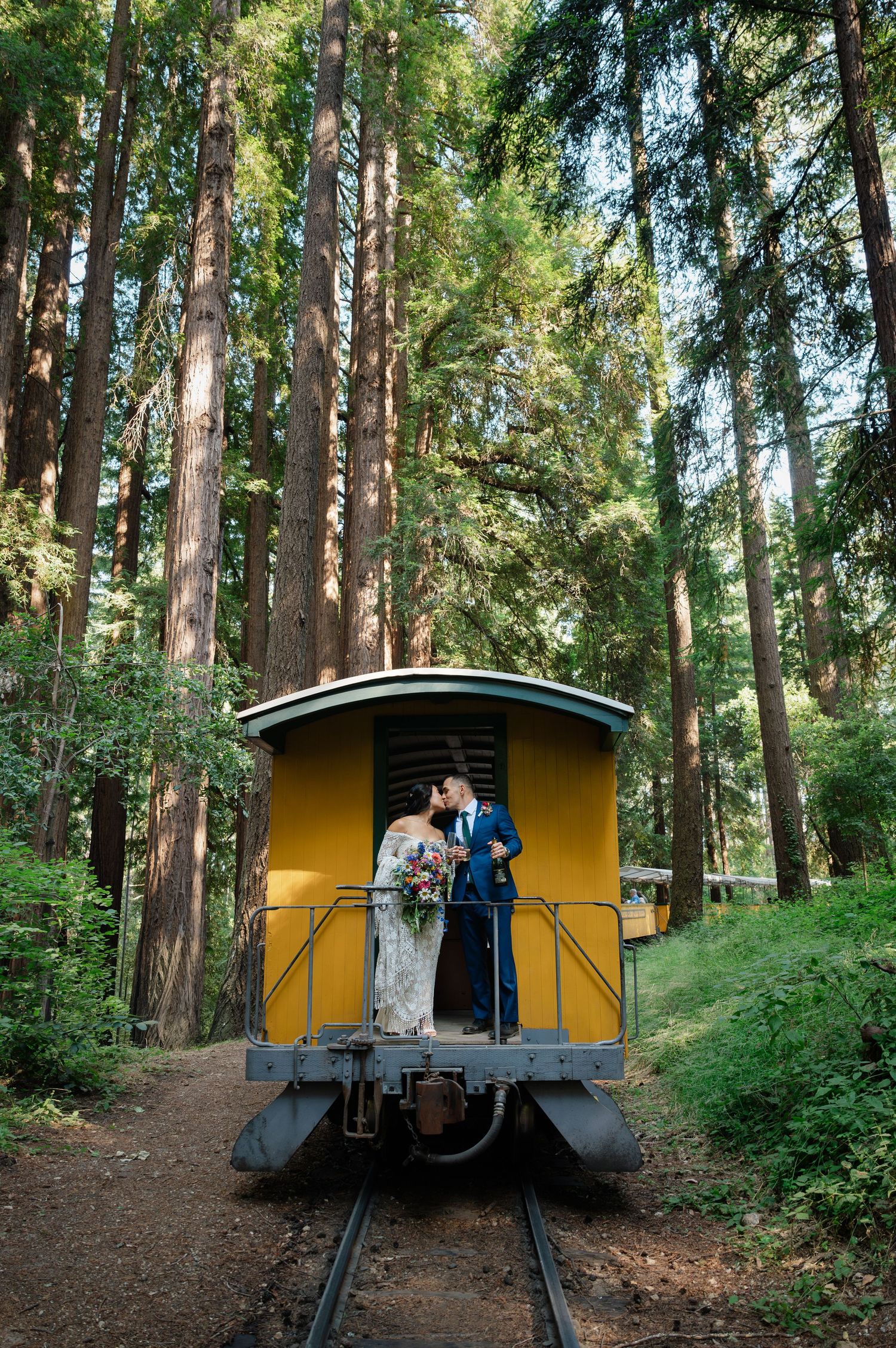 Bride and groom kissing on a yellow train car surrounded by tall redwood trees in a lush forest wedding photo.