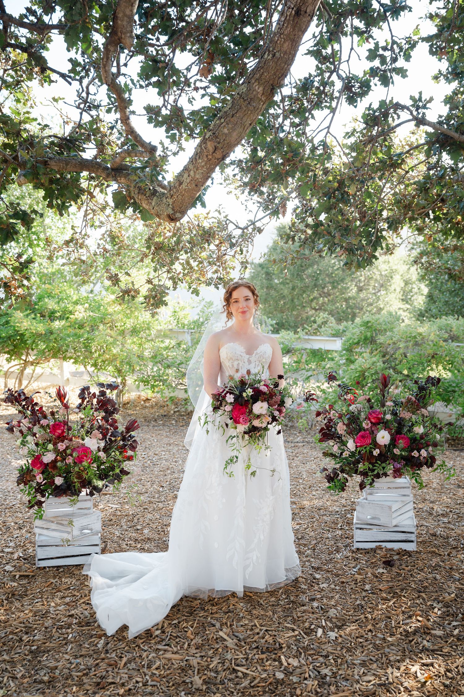 Elegant wedding scene with white crates and lush burgundy and white floral arrangements under a tree canopy.
