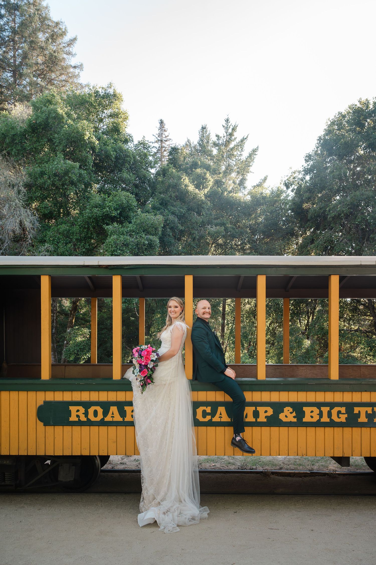 Bride and groom posing in front of Roaring Camp & Big Trees railroad car surrounded by lush green trees.
