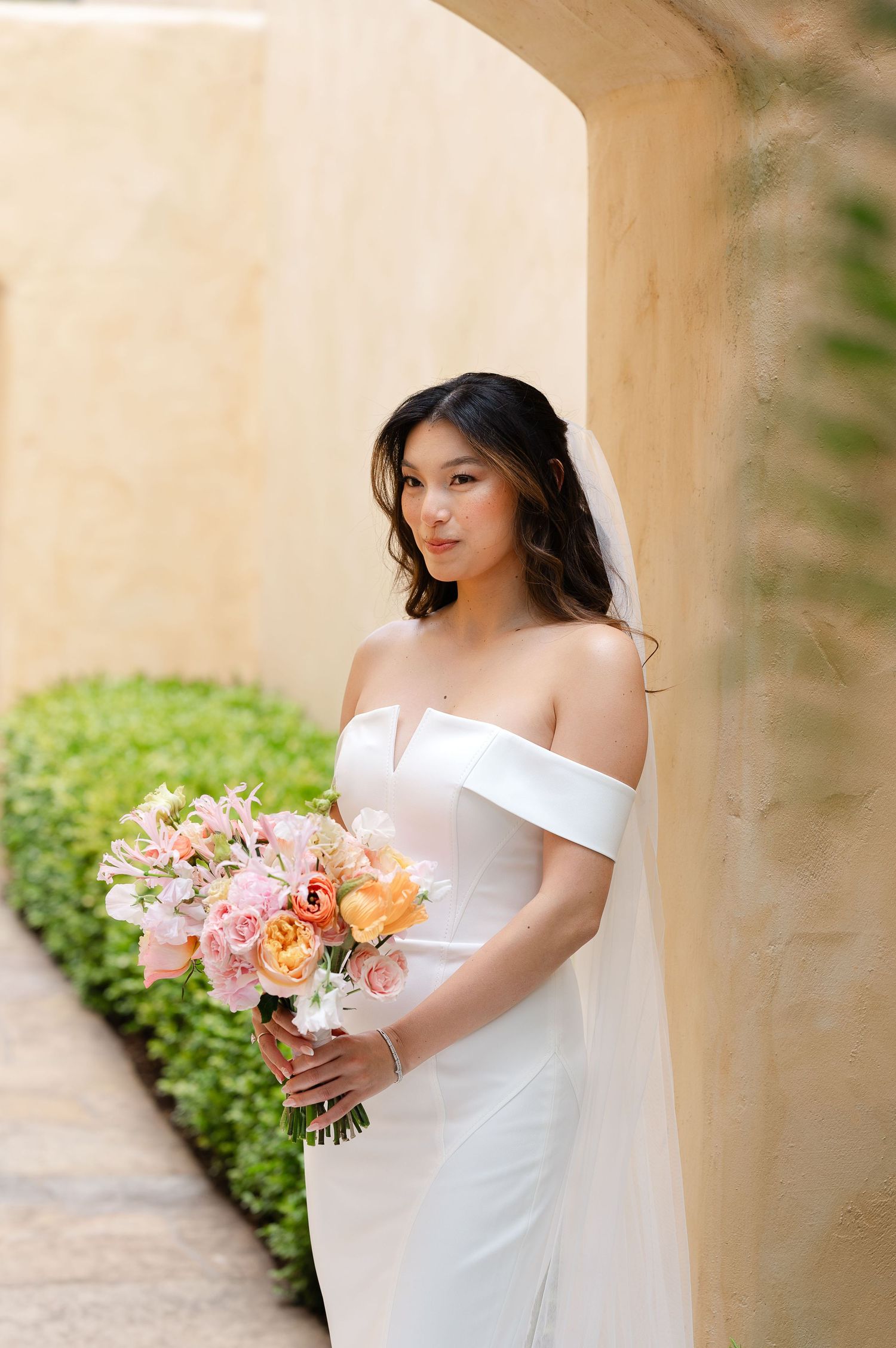 Elegant bridal portraits in white off-shoulder dress holding pink and peach floral bouquet against stone pillars.