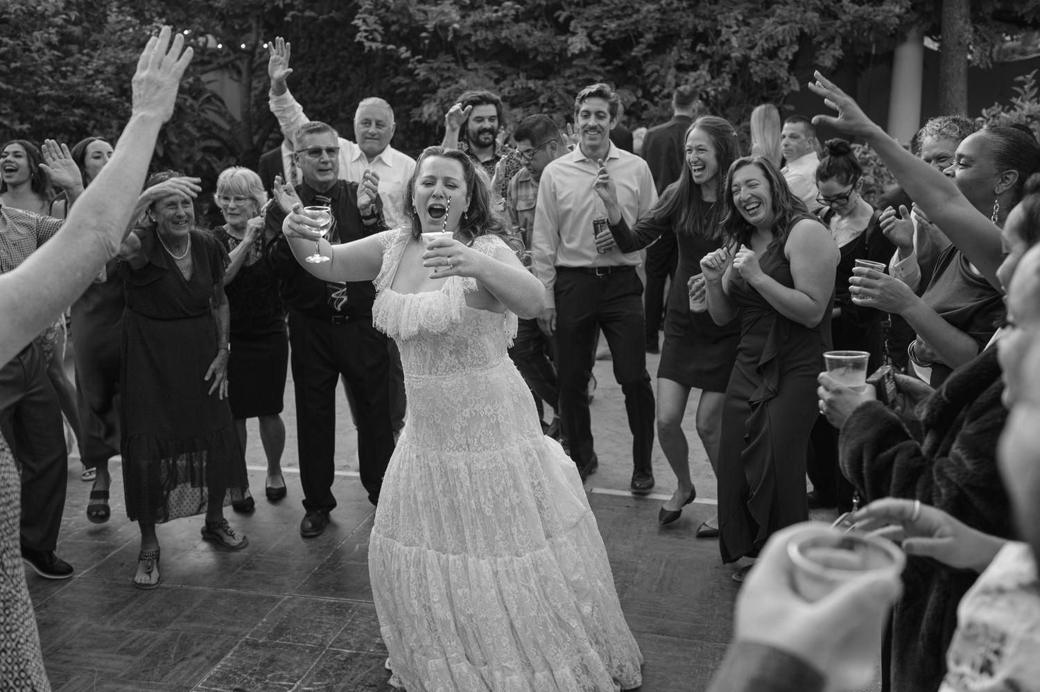 Bride dancing joyfully surrounded by clapping wedding guests in elegant attire at an outdoor reception.