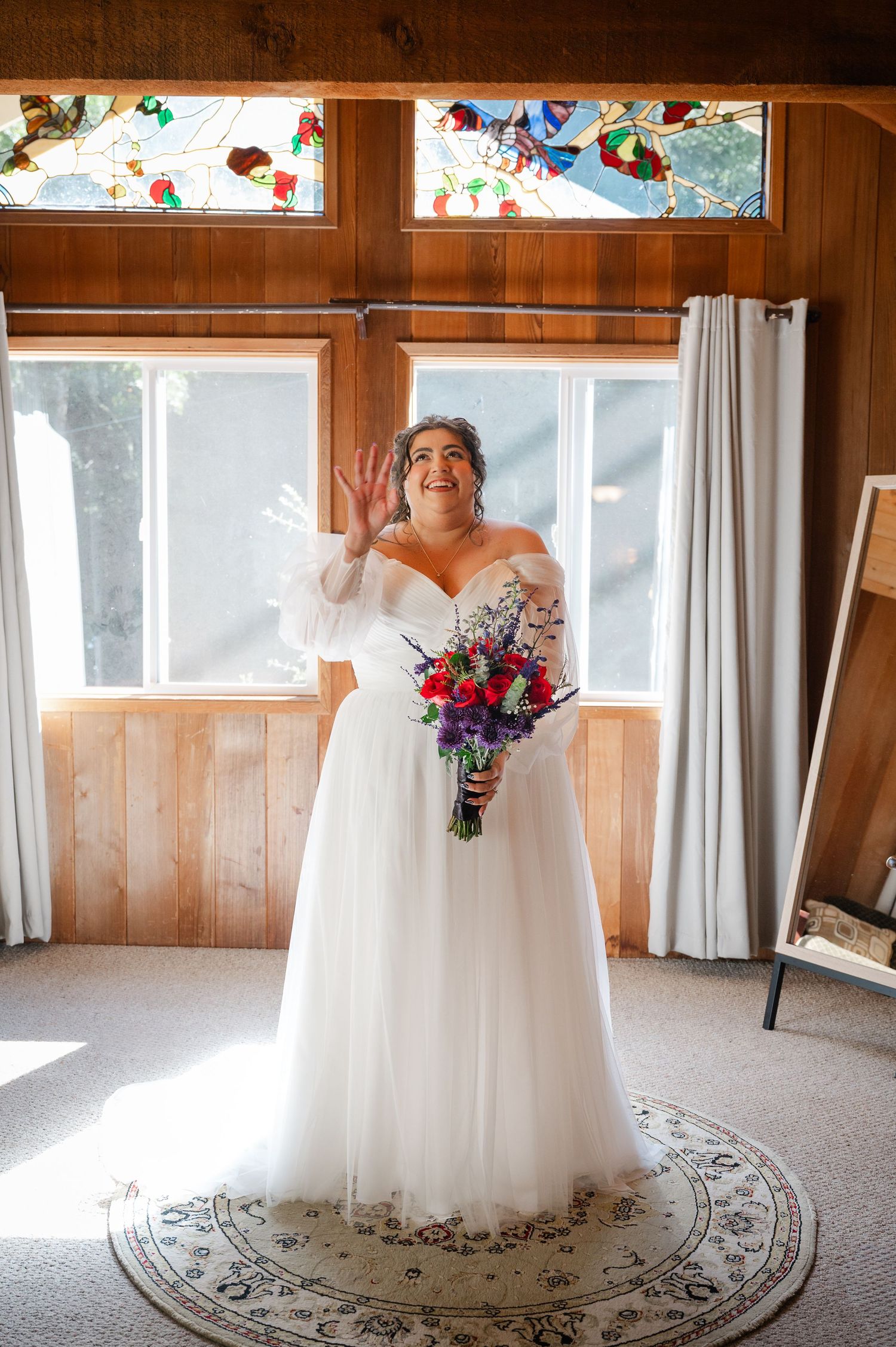 A bride in a white wedding dress stands on a patterned circular rug in a sunlit room with wood-paneled walls.