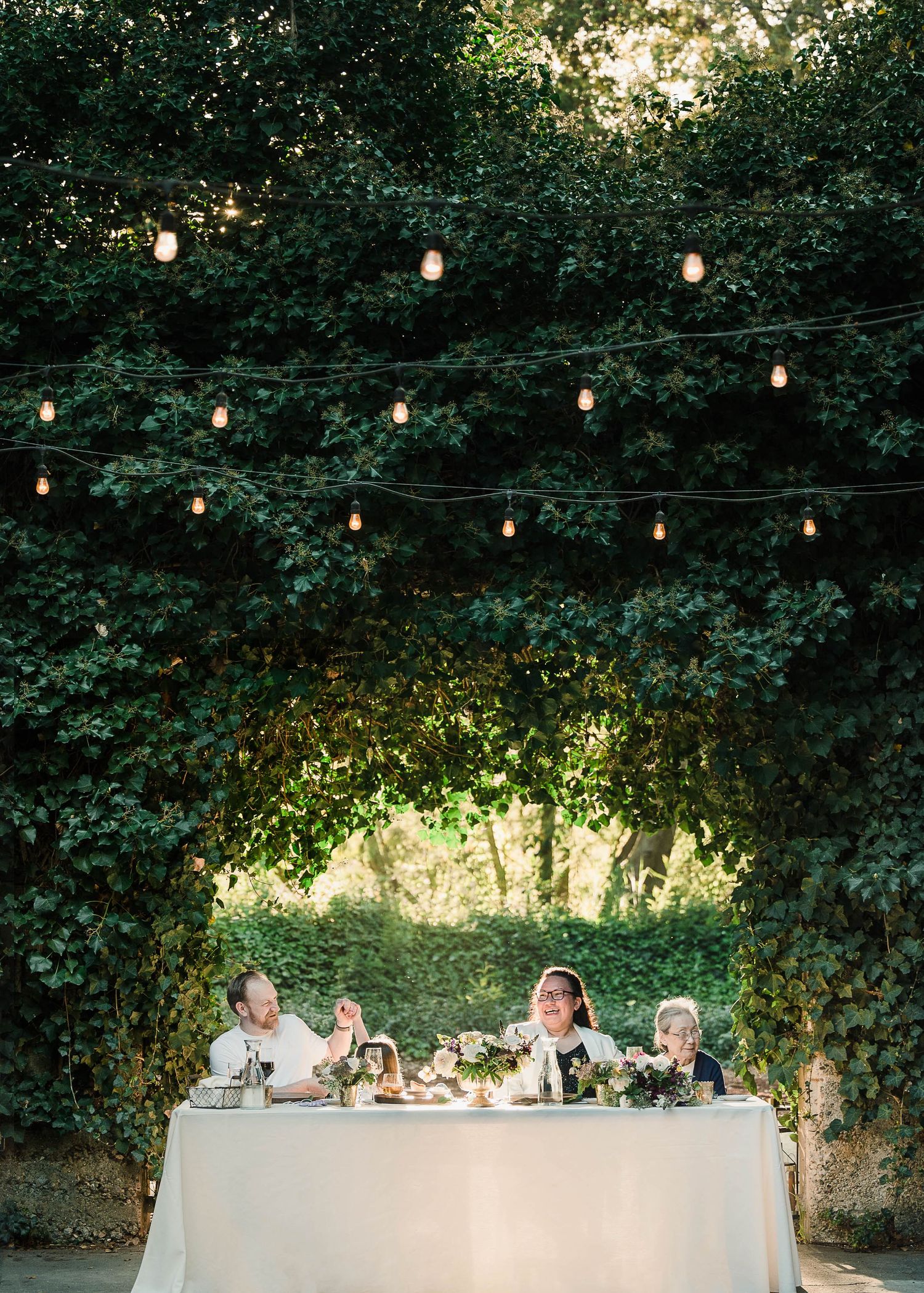 Outdoor wedding reception with string lights over a white draped sweetheart table, surrounded by lush green ivy walls.
