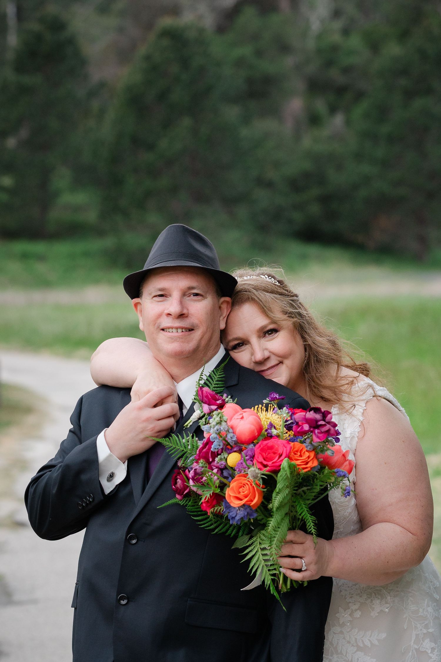 Smiling bride and groom pose outdoors, she holds colorful bouquet, he wears black hat and suit, lush green trees behind them.