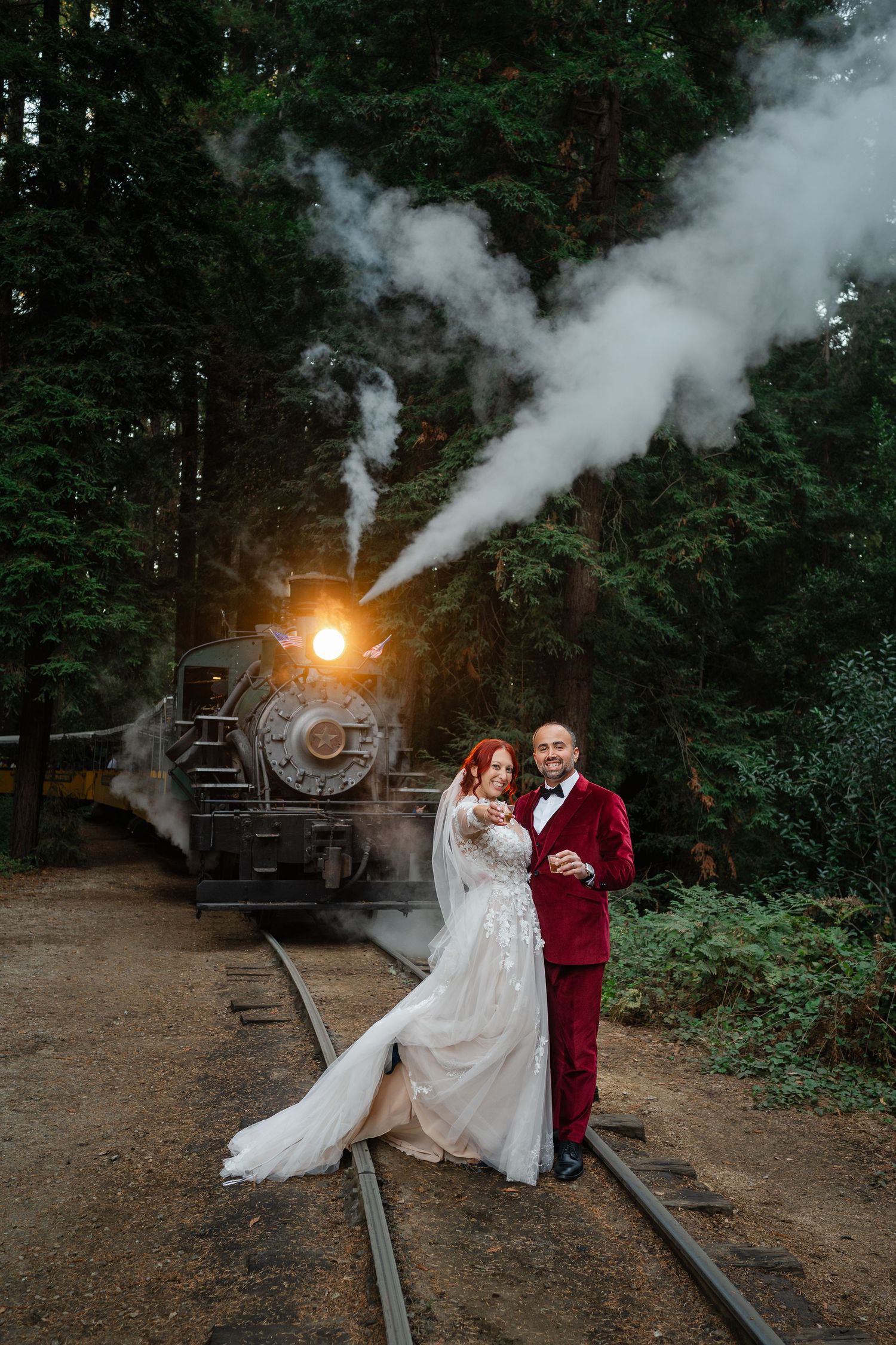 Bride and groom pose on railroad tracks before a vintage steam train in a lush forest wedding photo.