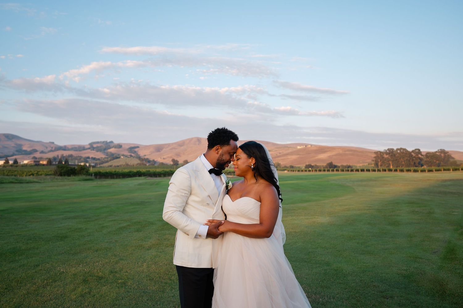 Bride and groom share a romantic moment at sunset in a lush green vineyard with rolling hills in the background.