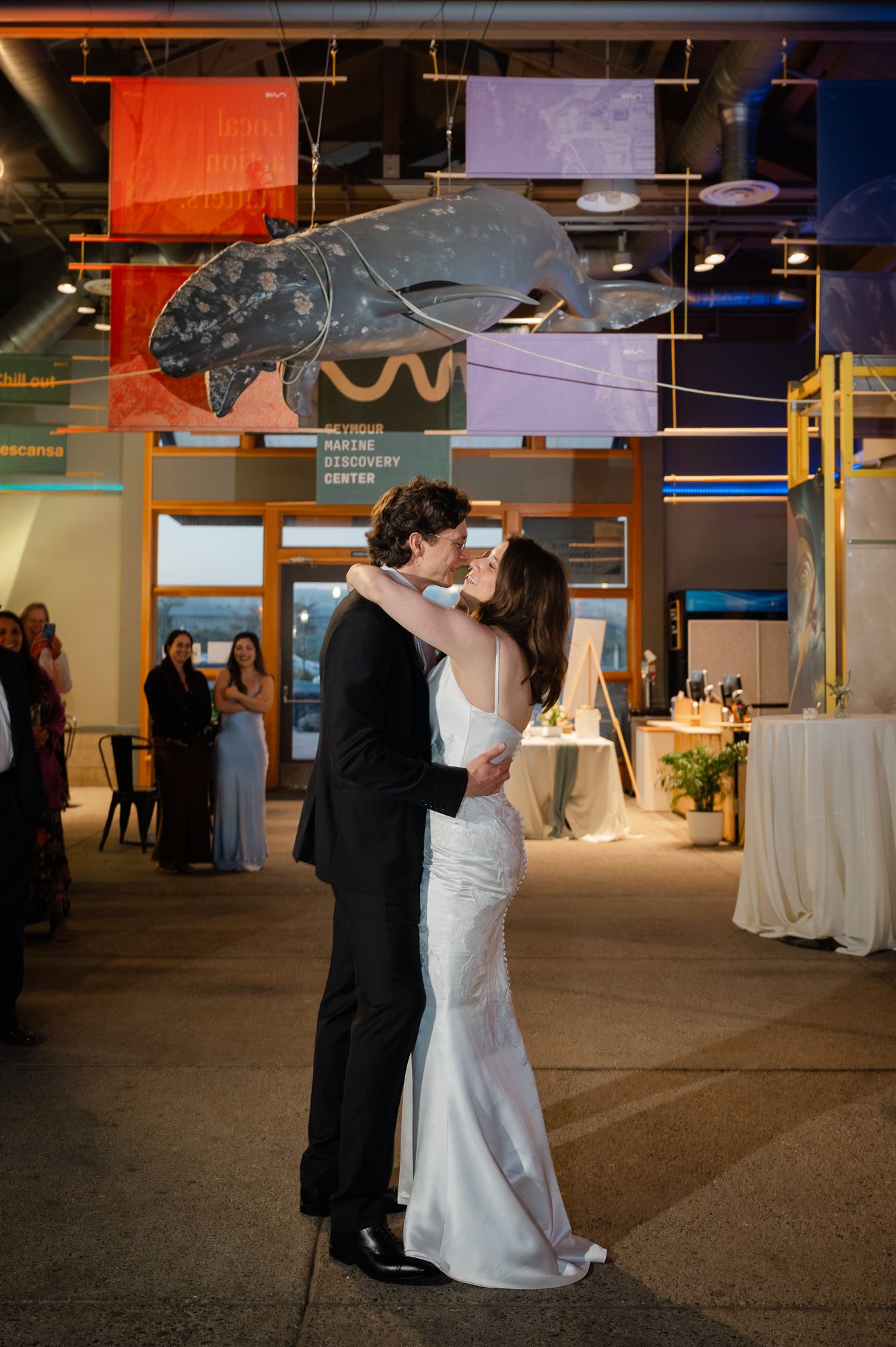 Bride and groom share first dance kiss at indoor wedding reception with colorful banners and guests in background.