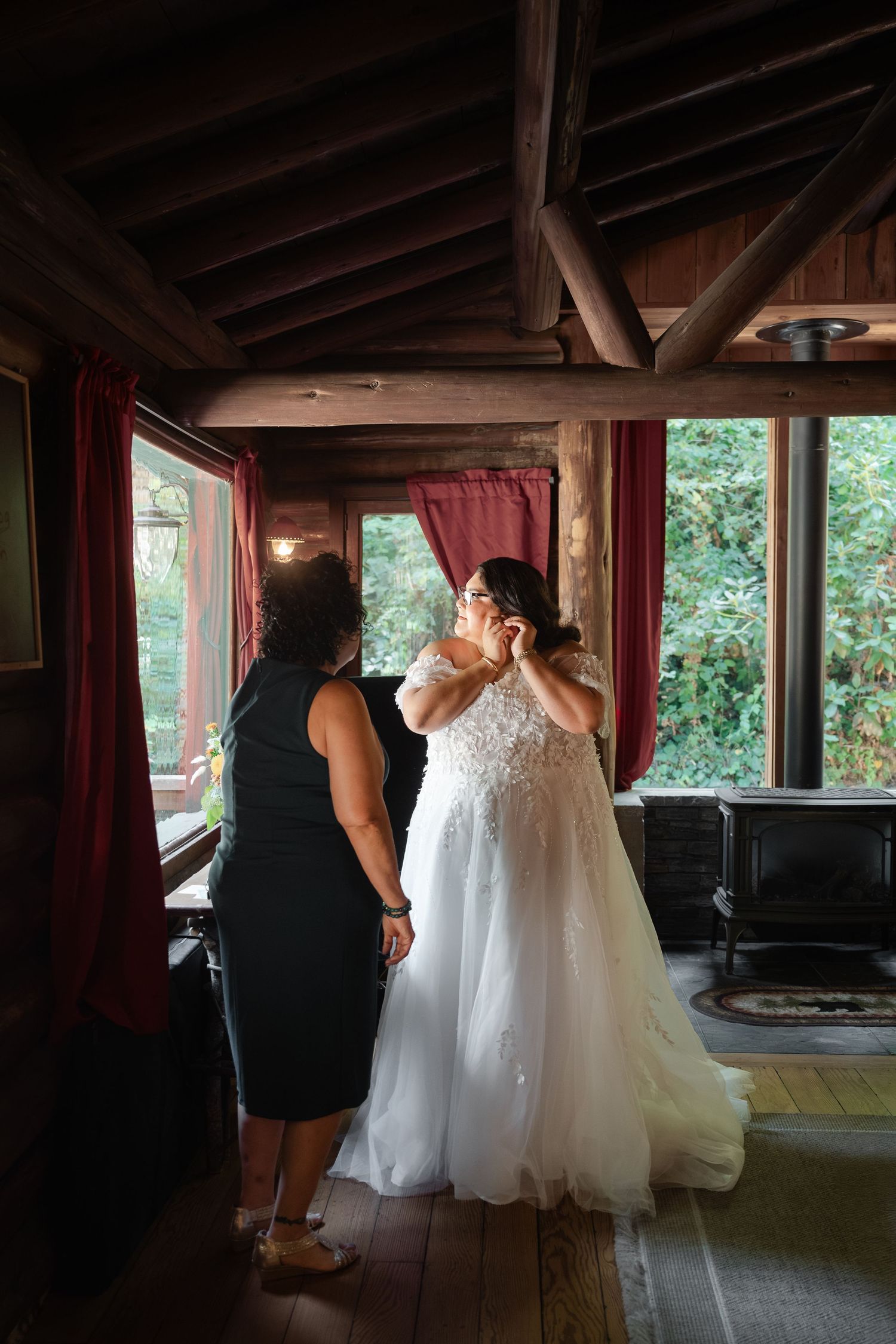 A wedding dress fitting session shows a seamstress making final adjustments in a rustic room with wooden beams and red curtains.