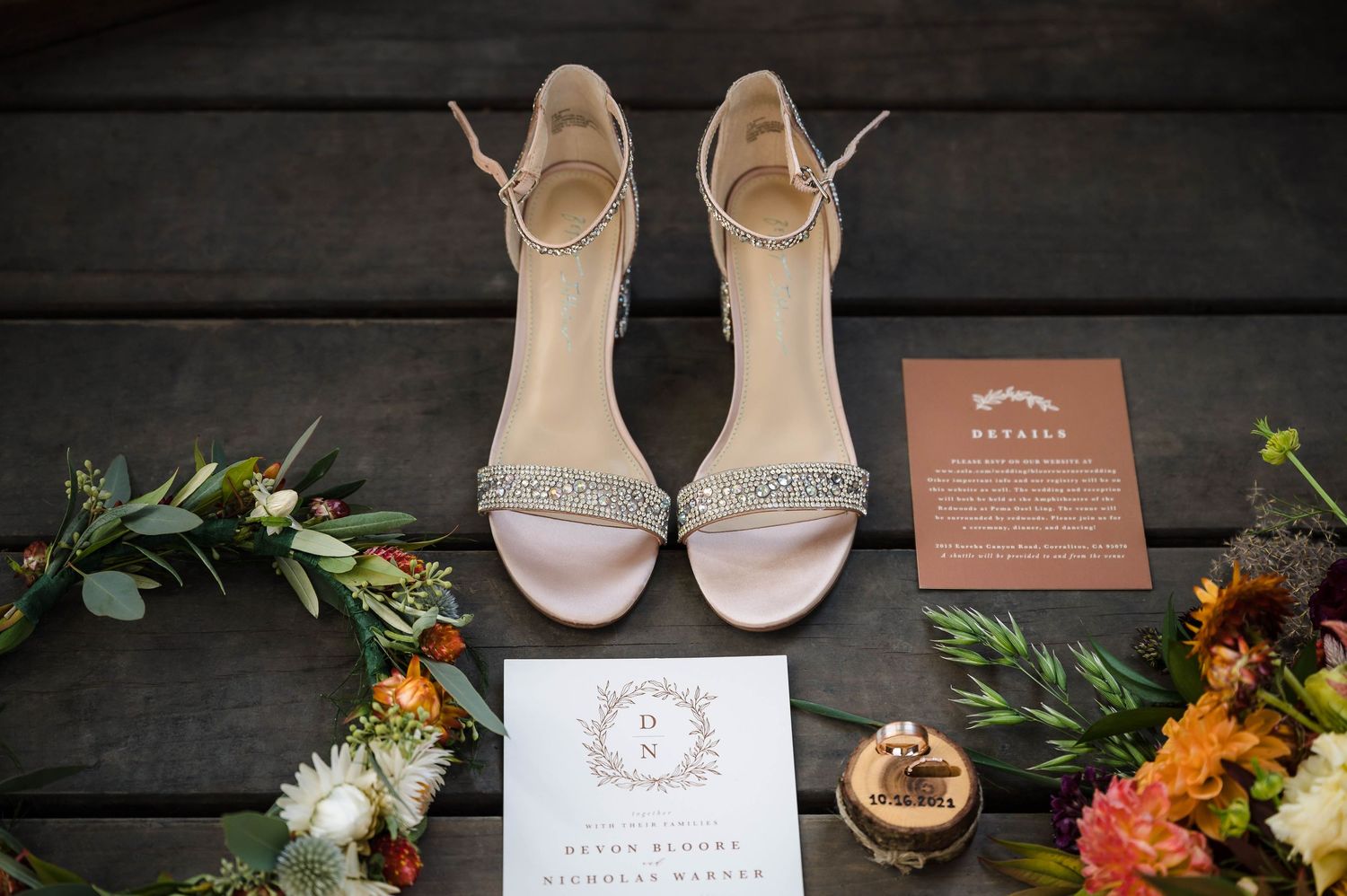 Elegant bridal shoes with invitation, wooden ring box, and colorful floral arrangements on dark wooden planks.
