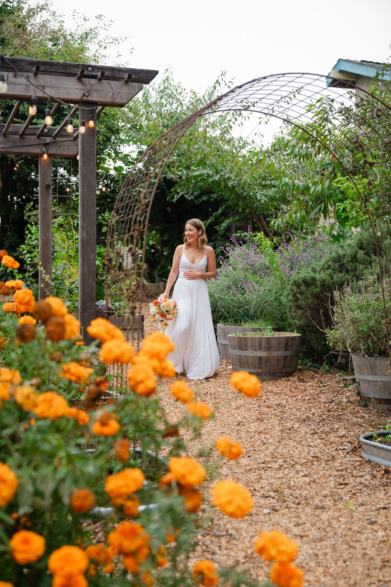 A garden path with orange marigolds and purple flowers leads to a wooden pergola in a romantic outdoor setting.