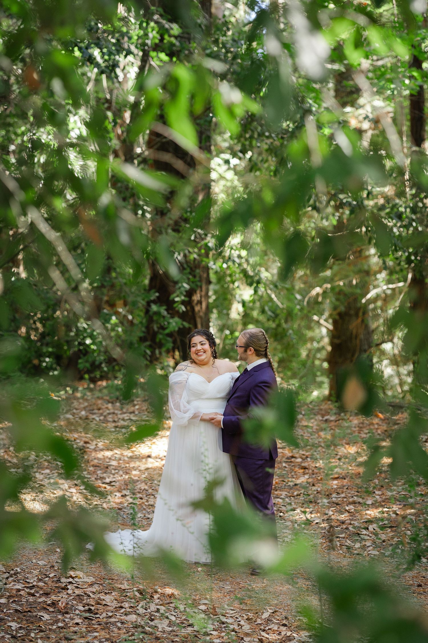 Bride and groom share a romantic moment on a forest path, surrounded by lush green trees and dappled sunlight.