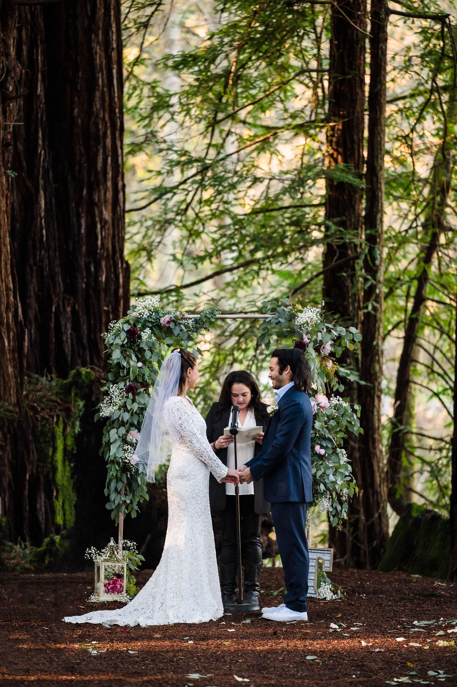 Couple exchanging vows under floral arch in redwood forest wedding ceremony with officiant.