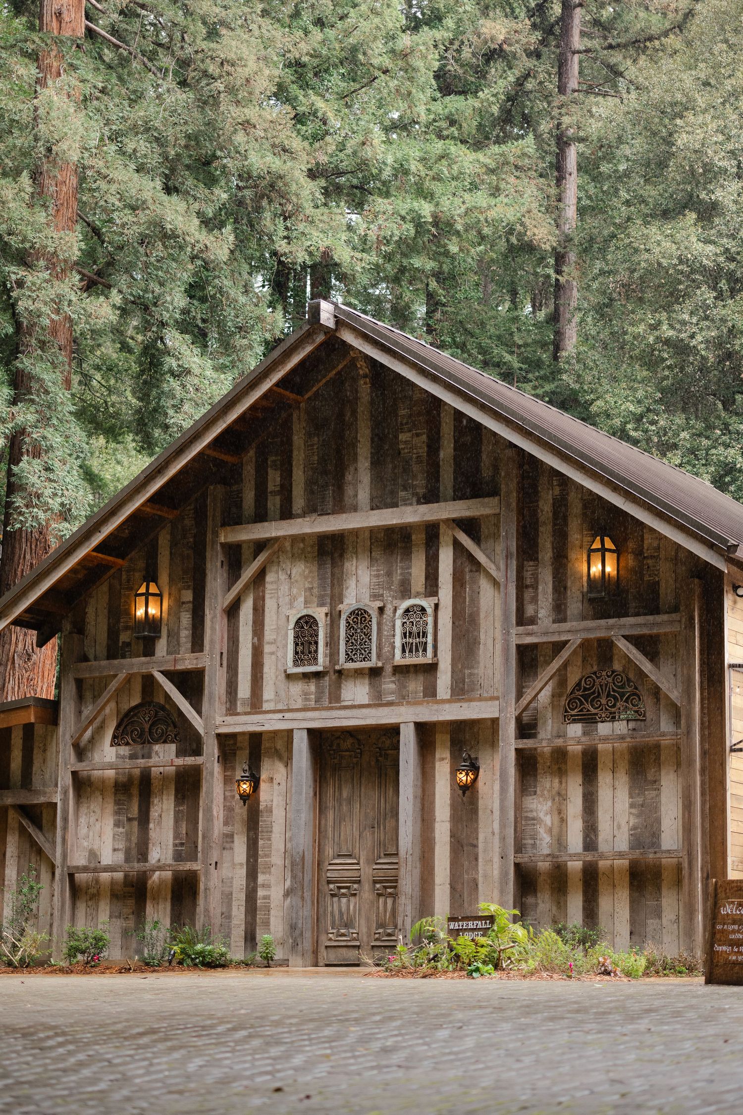 Rustic wooden barn with ornate doors, lantern sconces, and decorative windows nestled among tall evergreen trees.