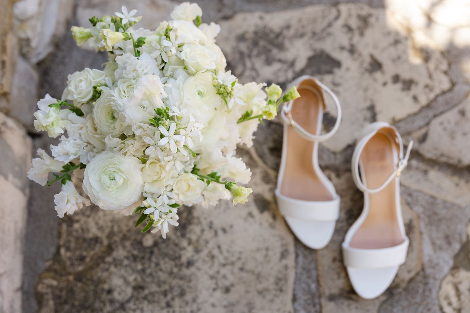 White bridal shoes and delicate floral bouquet with ranunculus and lilies arranged on rustic stone surface.