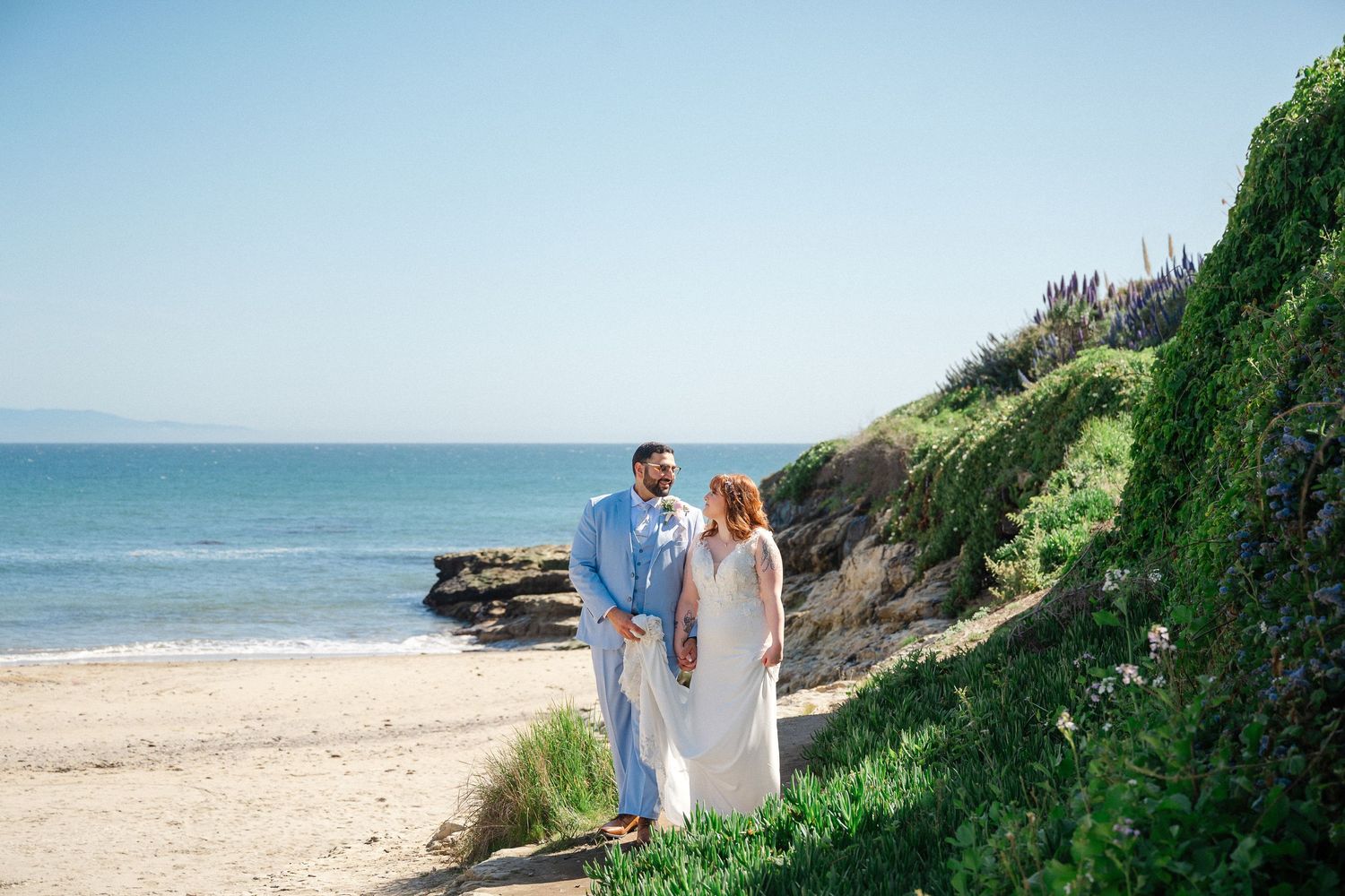 Couple in wedding attire holding hands on a scenic beach path with green cliffs and ocean views on a sunny day.