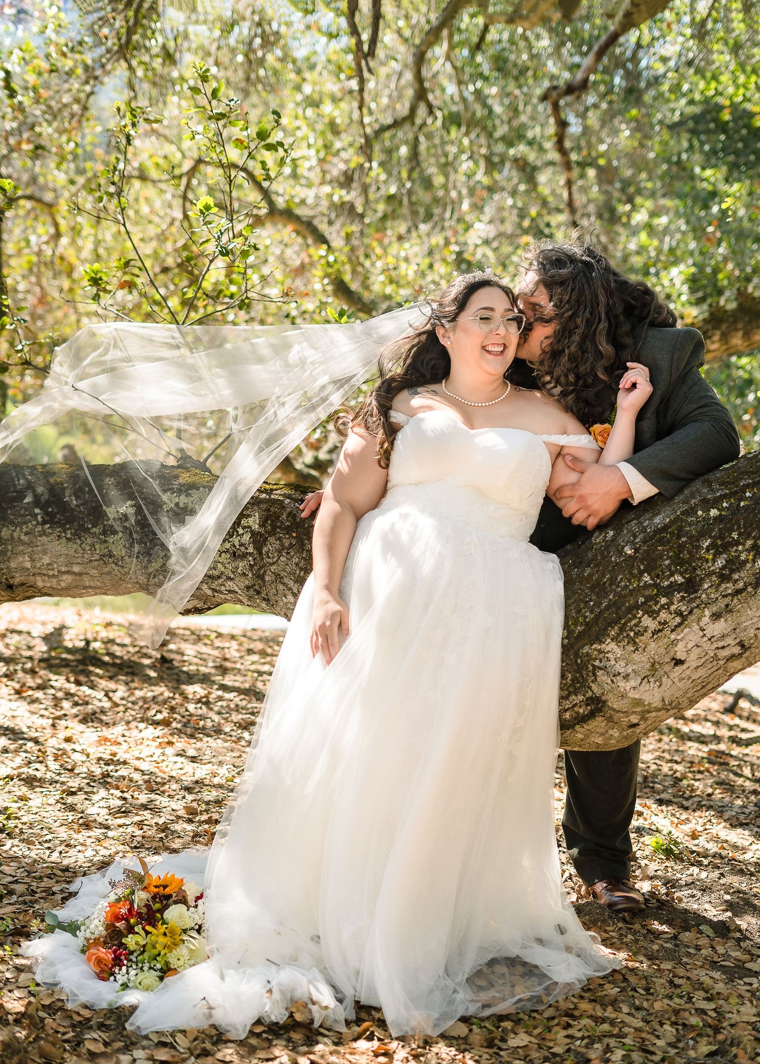 A romantic wedding couple poses together on fallen logs in a sunlit forest setting with autumn flowers.