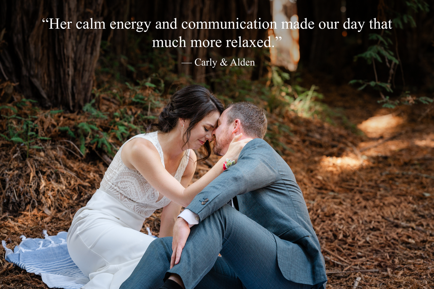 Couple sitting together on forest floor in wedding attire, bride in white gown and groom in gray suit, sharing intimate moment.