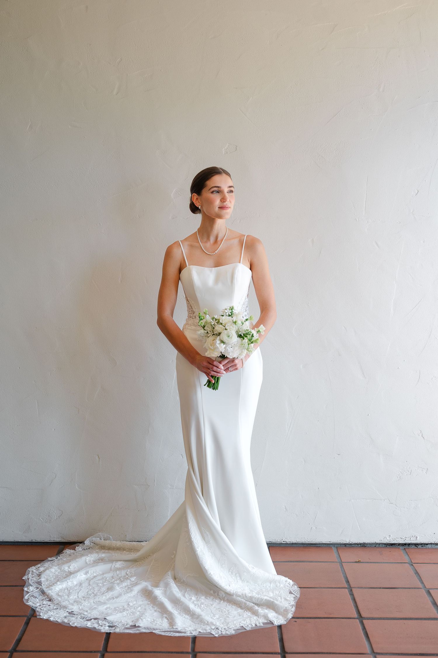 A person in a flowing white wedding dress poses elegantly on terracotta tiles against a neutral wall.