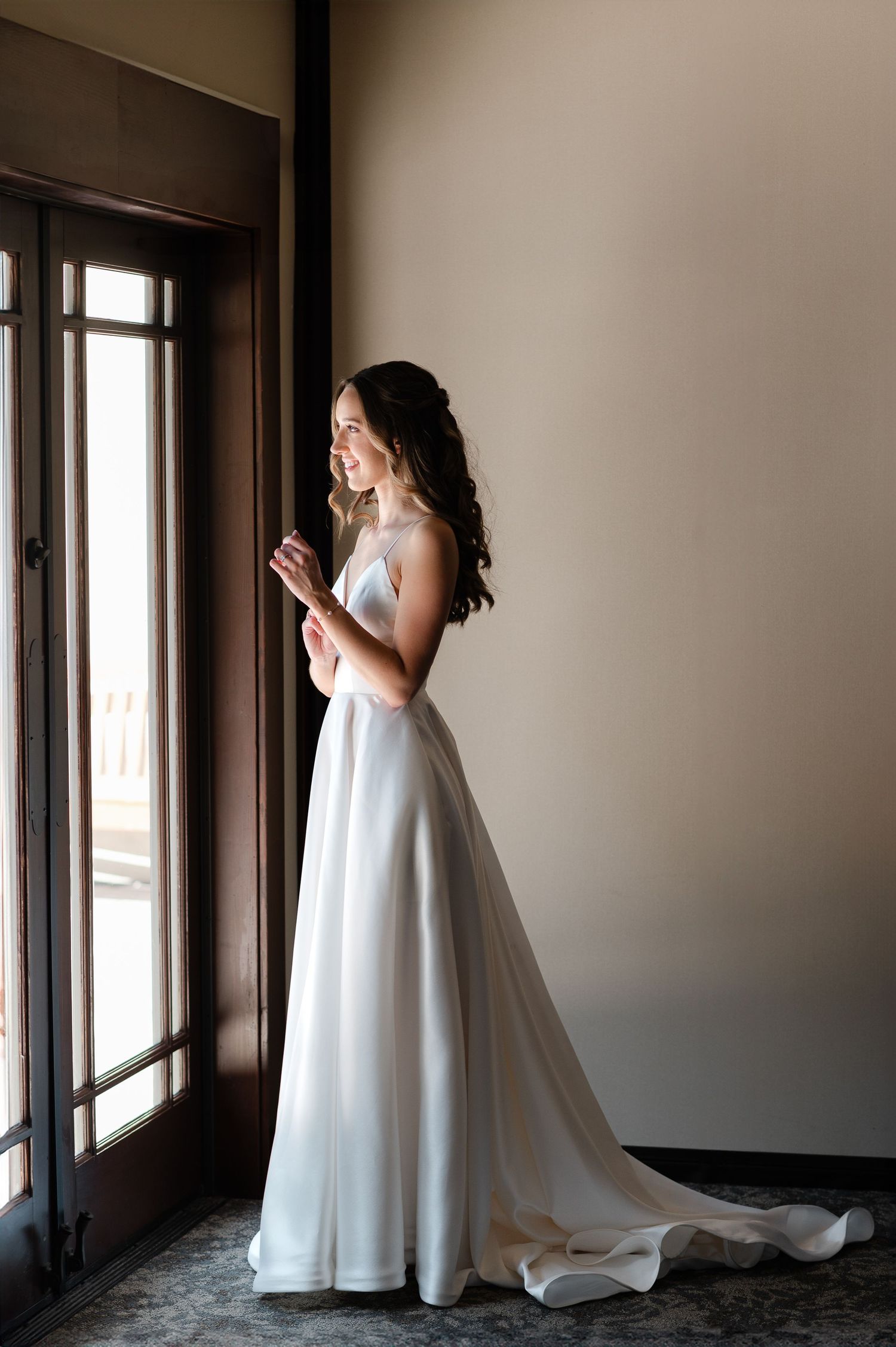 A bride in a flowing white wedding gown stands elegantly by large windows in a series of connected photos.