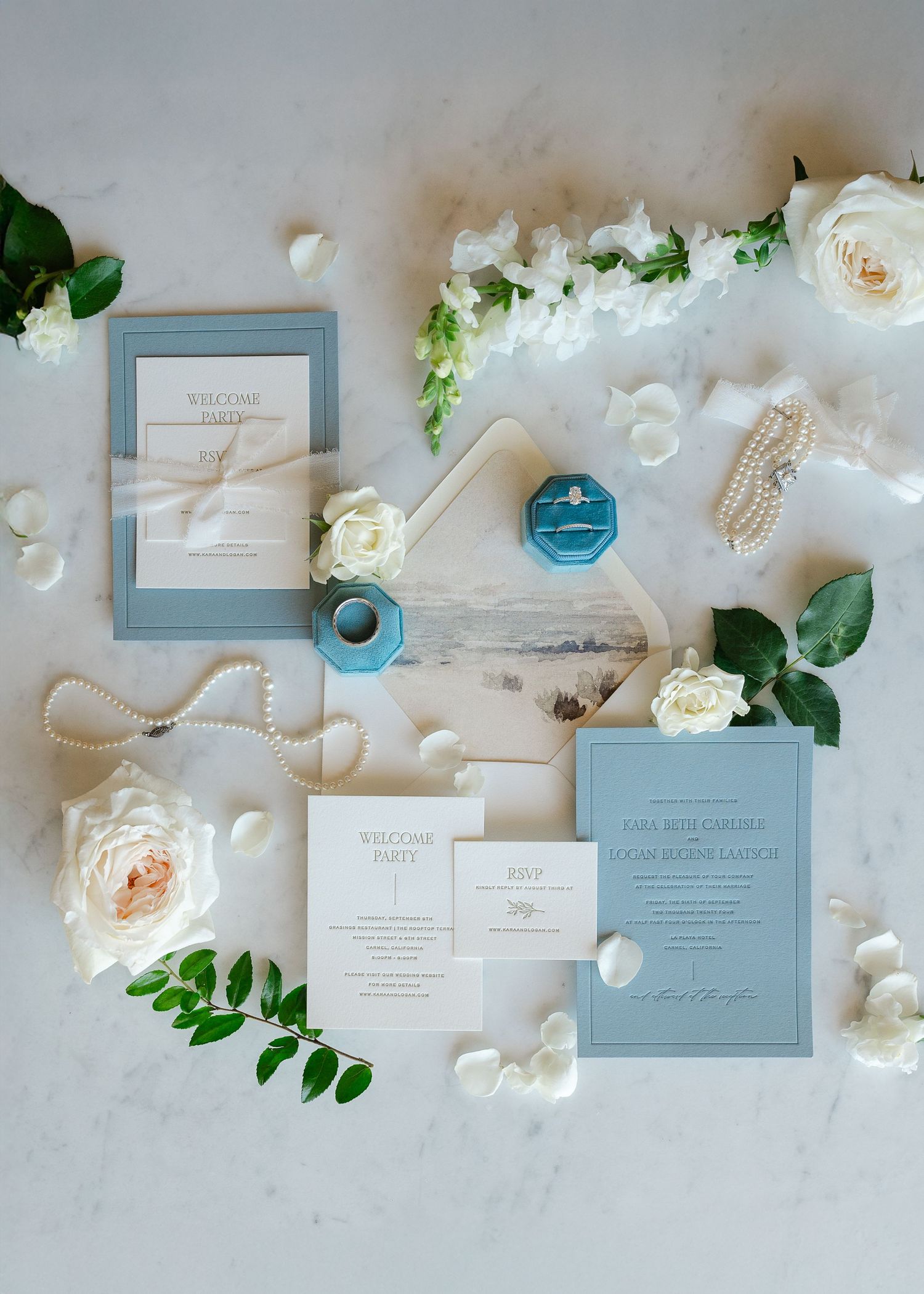 Elegant wedding stationery suite in dusty blue and white with fresh roses and greenery arranged on marble surface.