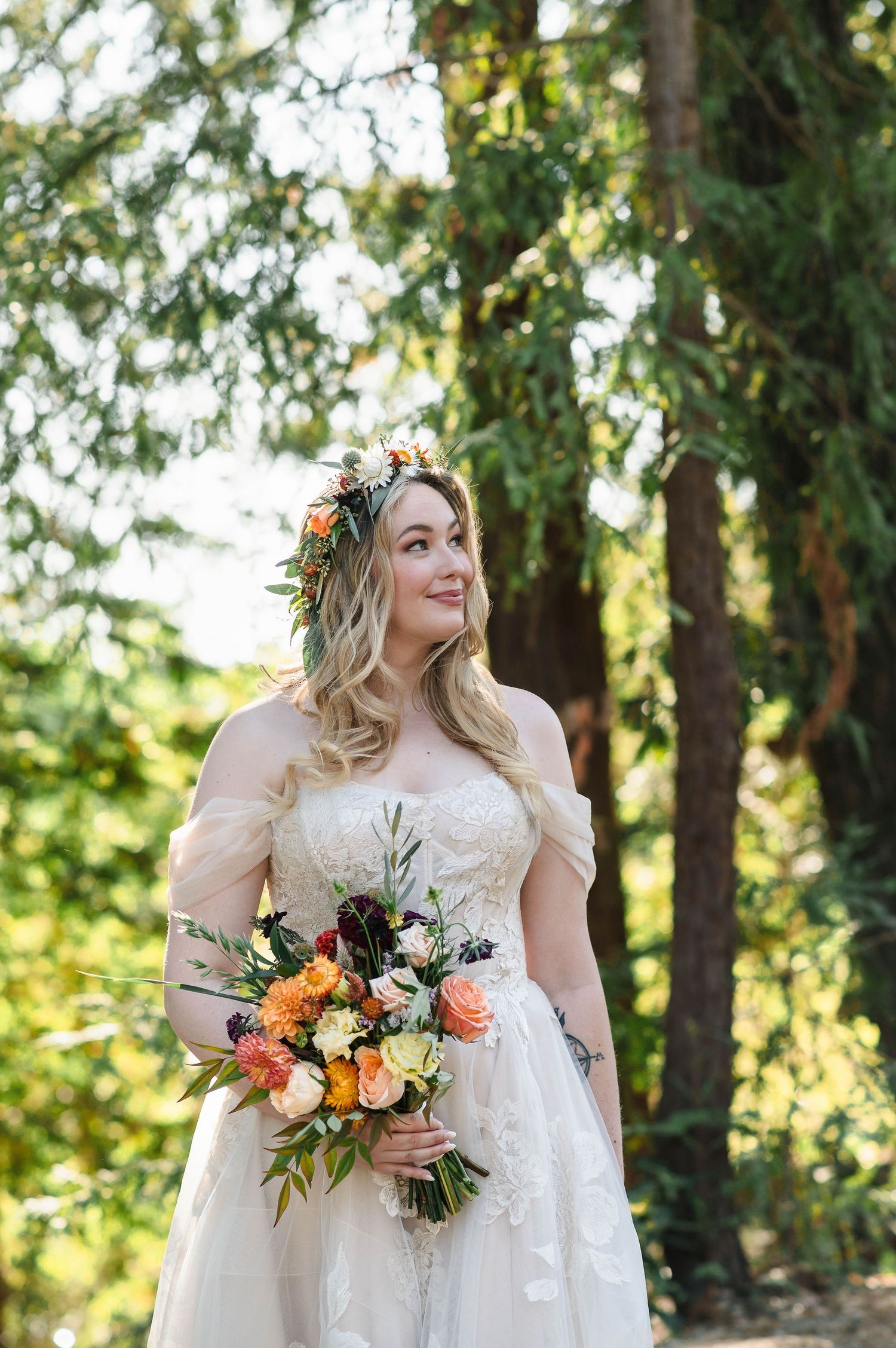 A bride in a lace wedding dress holds a vibrant orange and white floral bouquet in a sunlit forest setting.