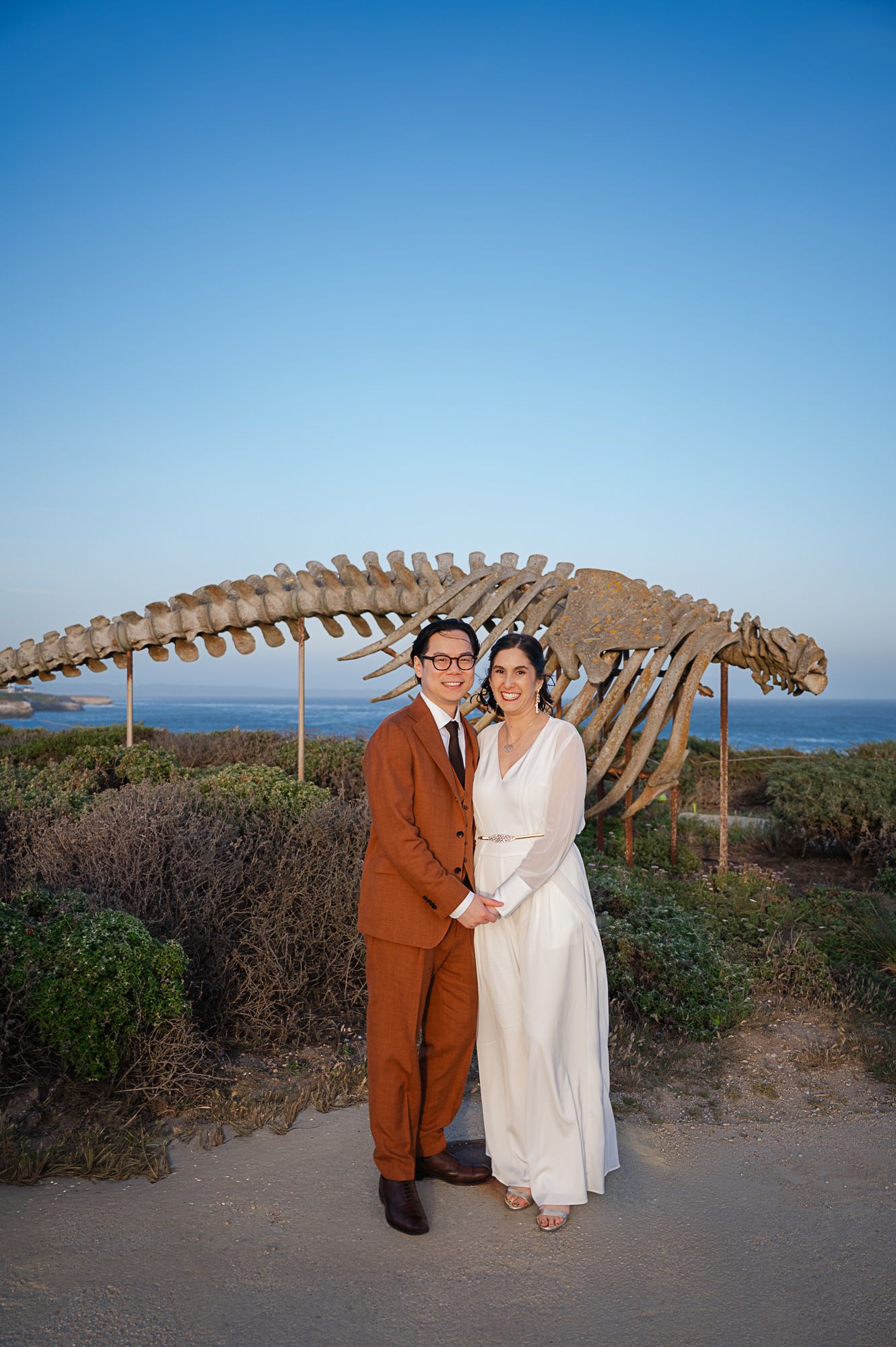 Couple in wedding attire poses before a whale skeleton sculpture with ocean views at sunset.