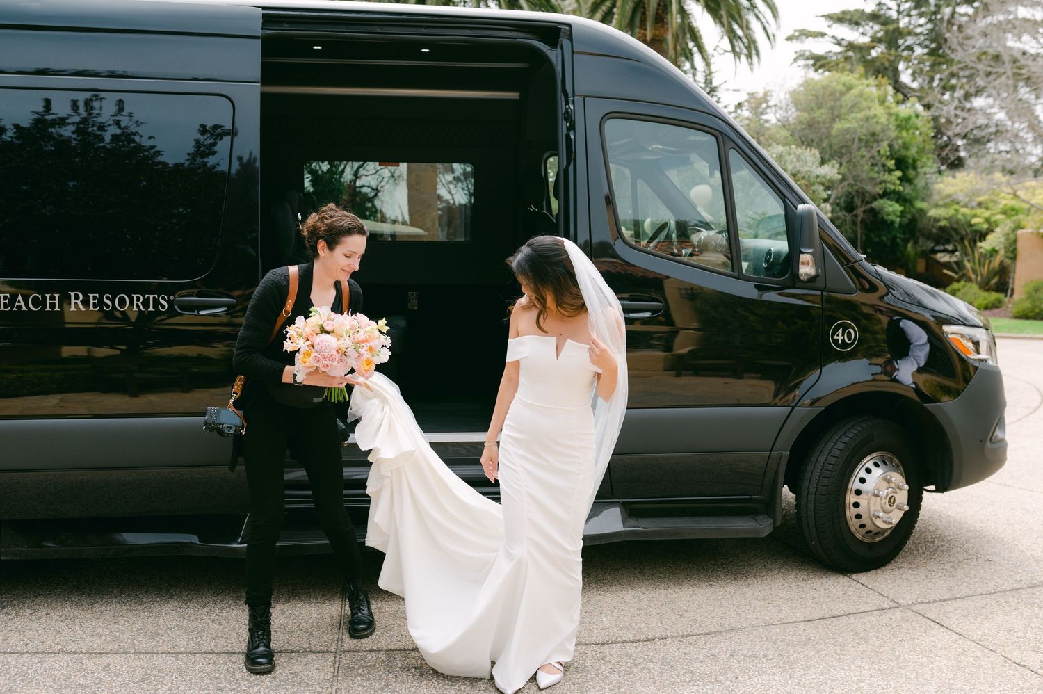 Bride in white gown holds pink bouquet beside groom near black luxury van at elegant wedding venue.