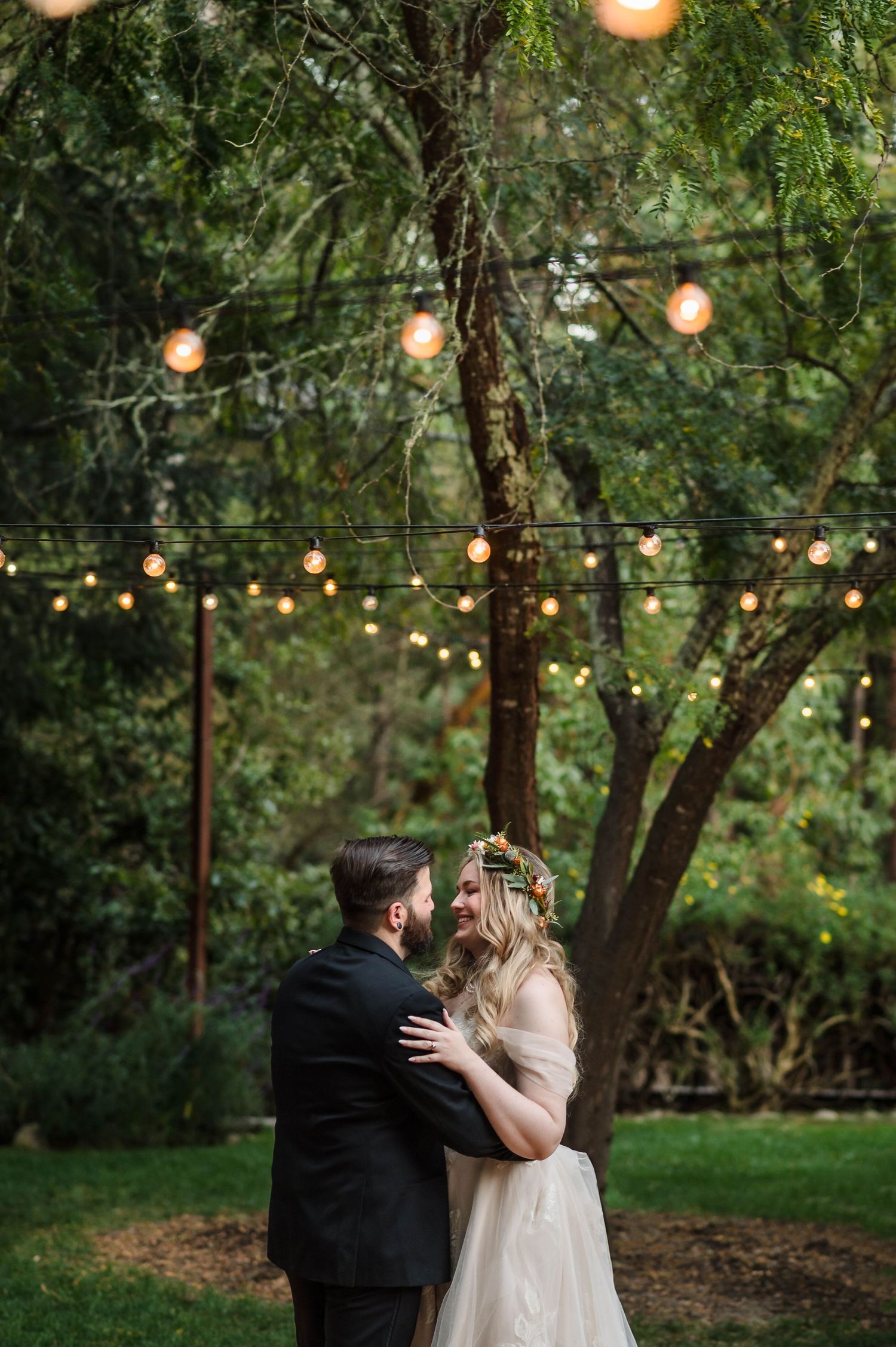 Couple embraces under string lights in lush garden at twilight wedding, bride in floral crown and groom in black suit.
